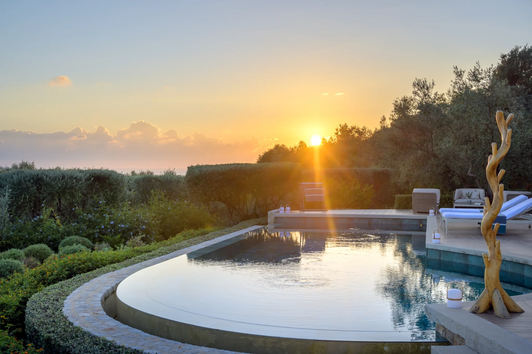Setting sun above olive trees reflected in a curved swimming pool, with sun loungers and a sculptural timber feature on the terrace at The Olive Retreat near Pylos.