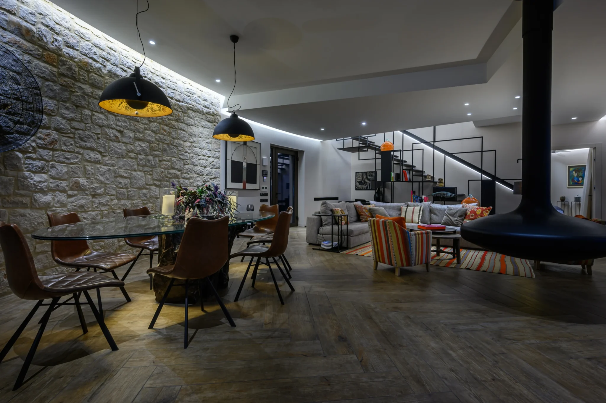 Glass dining table and leather chairs beneath black pendant lights beside a stone wall, with a suspended fireplace and living room beyond at The Olive Retreat near Pylos.
