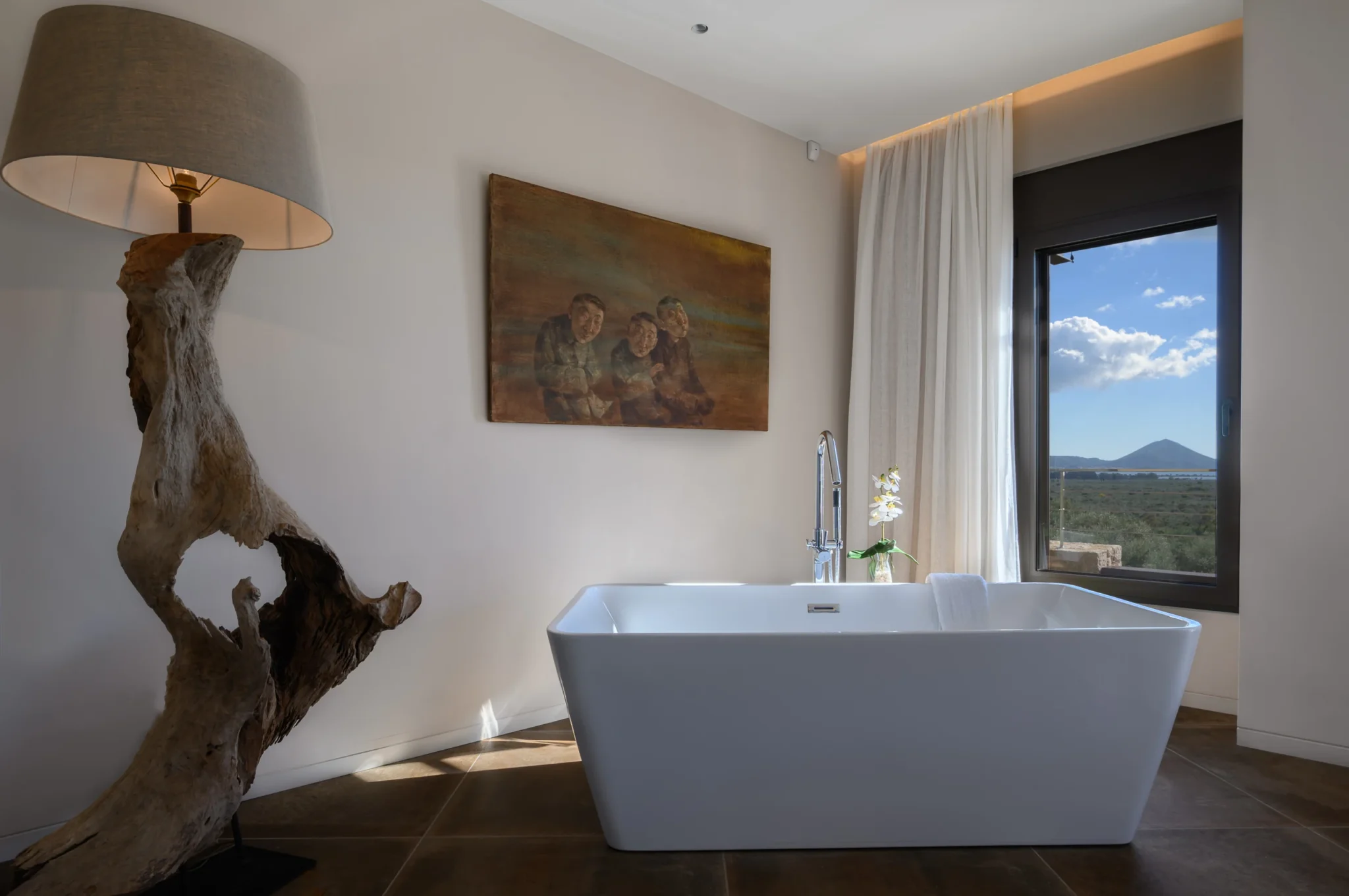 Freestanding white bath beside a landscape-view window, with a sculptural timber floor lamp and a figurative painting at The Olive Retreat near Pylos.