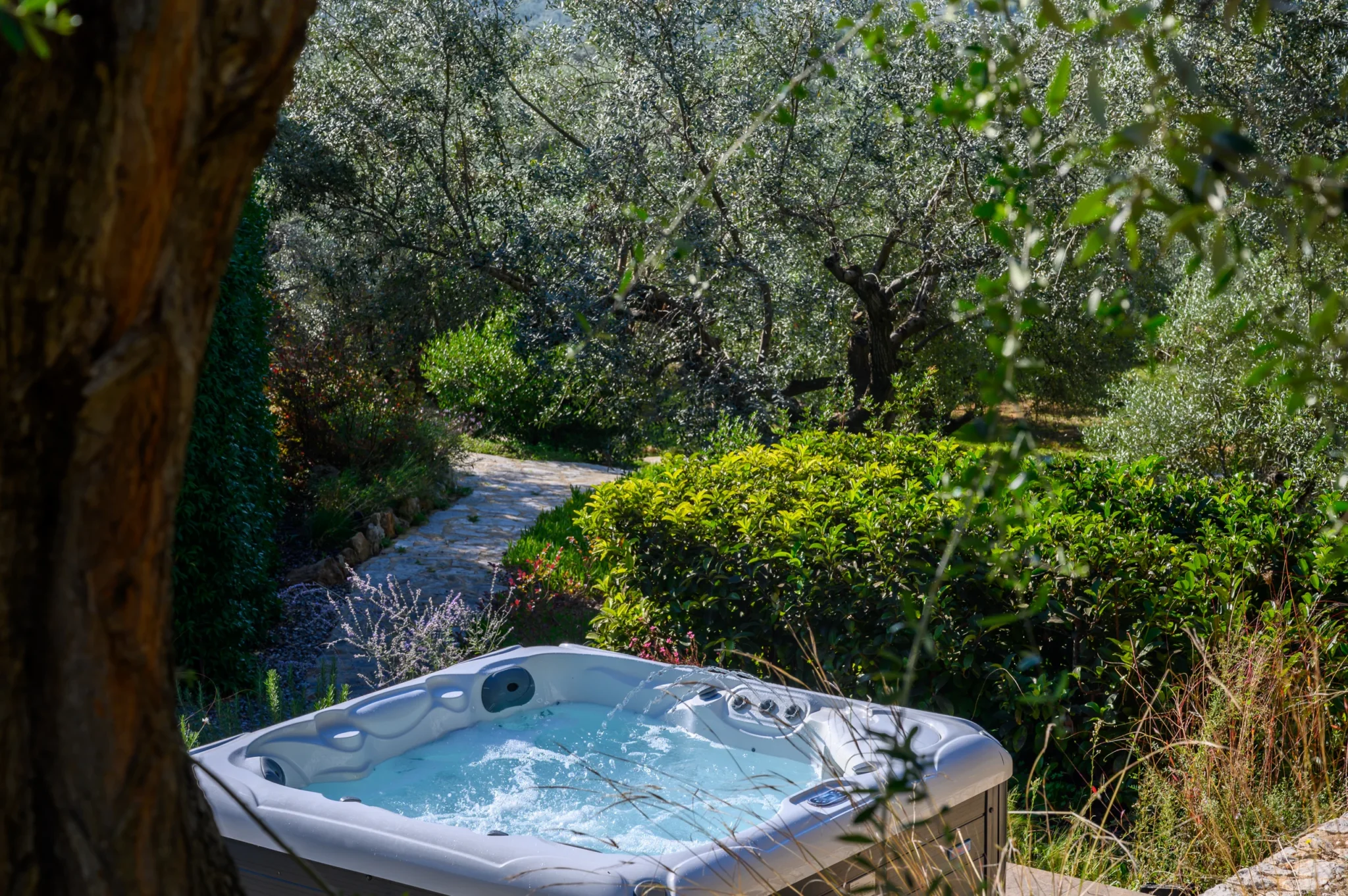 Outdoor hot tub with flowing water jets among mature olive trees and dense garden planting beside a stone path at The Olive Retreat near Pylos.