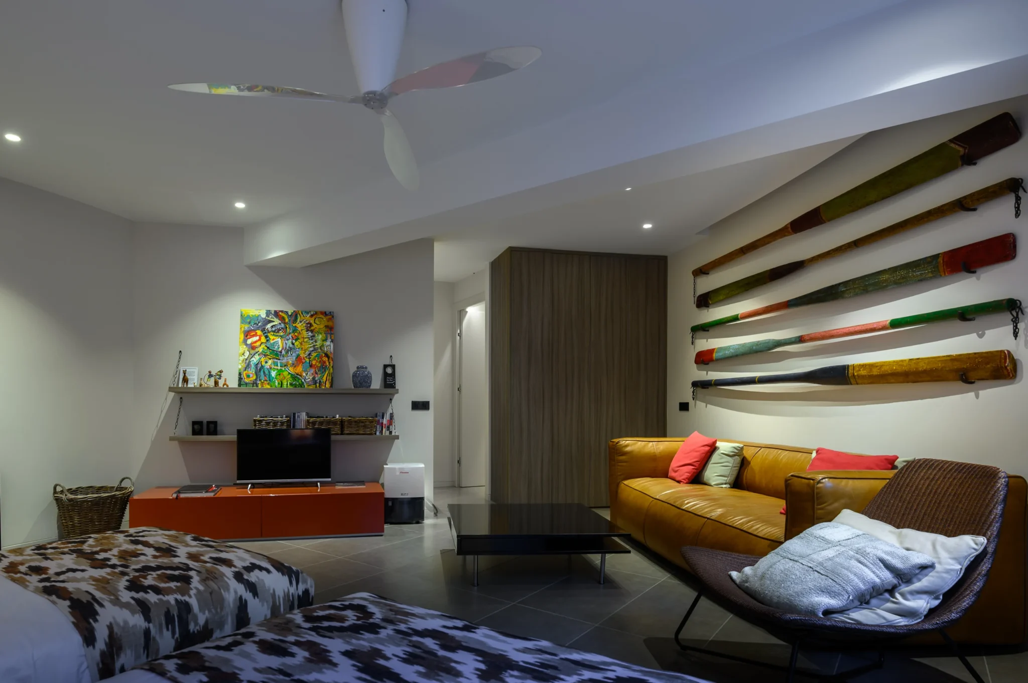 Guest room with twin beds, a tan leather sofa, red cushions, colourful wall-mounted wooden oars and an orange television console at The Olive Retreat near Pylos.