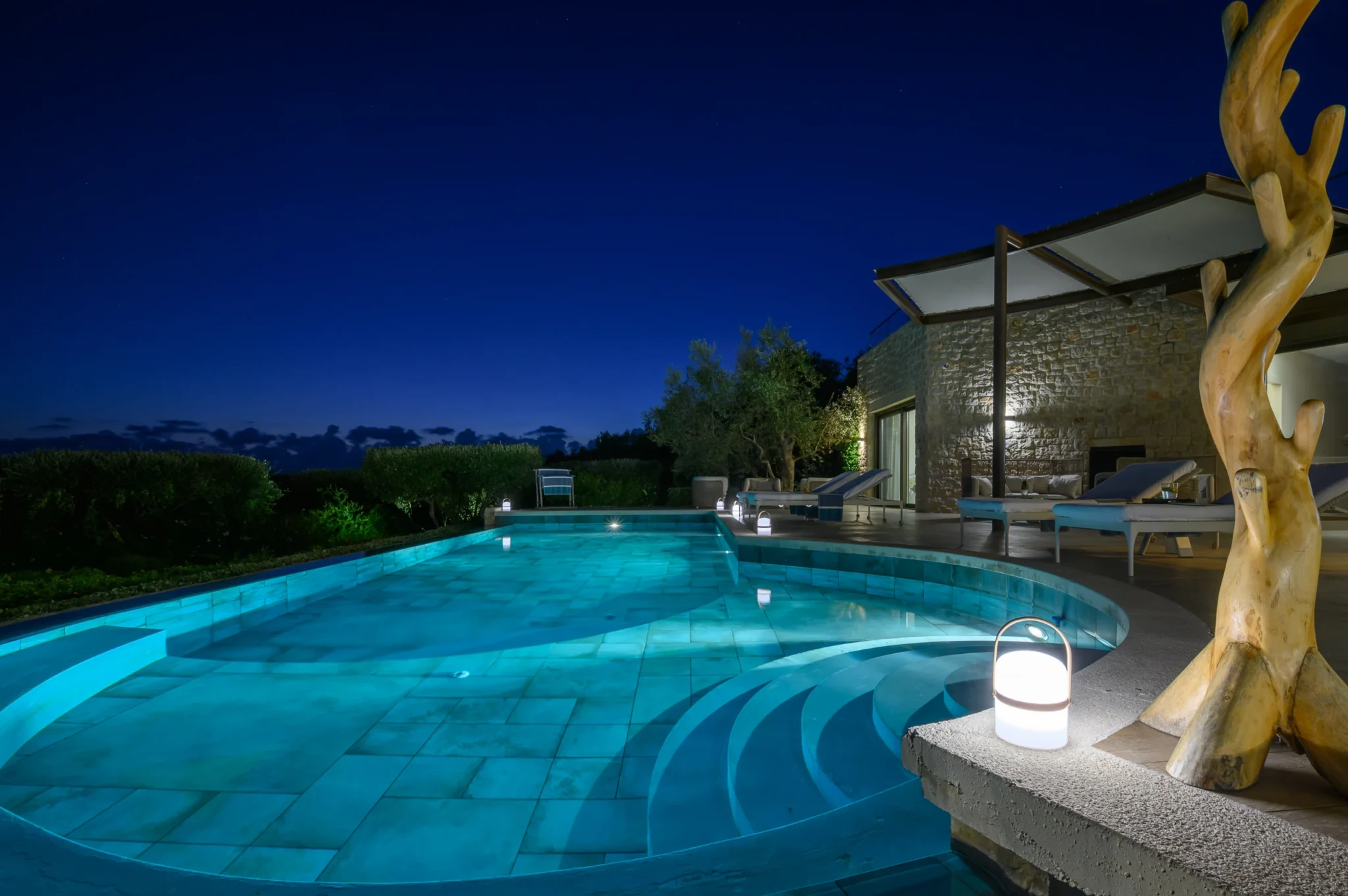 Turquoise illuminated swimming pool with curved underwater steps, a terrace lantern and sculptural timber feature beside the stone facade at night at The Olive Retreat near Pylos.