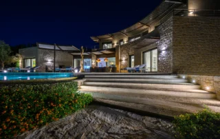 Illuminated stone facade with pergolas, broad curved steps and a covered dining terrace beside the swimming pool and planted garden at night at The Olive Retreat near Pylos.