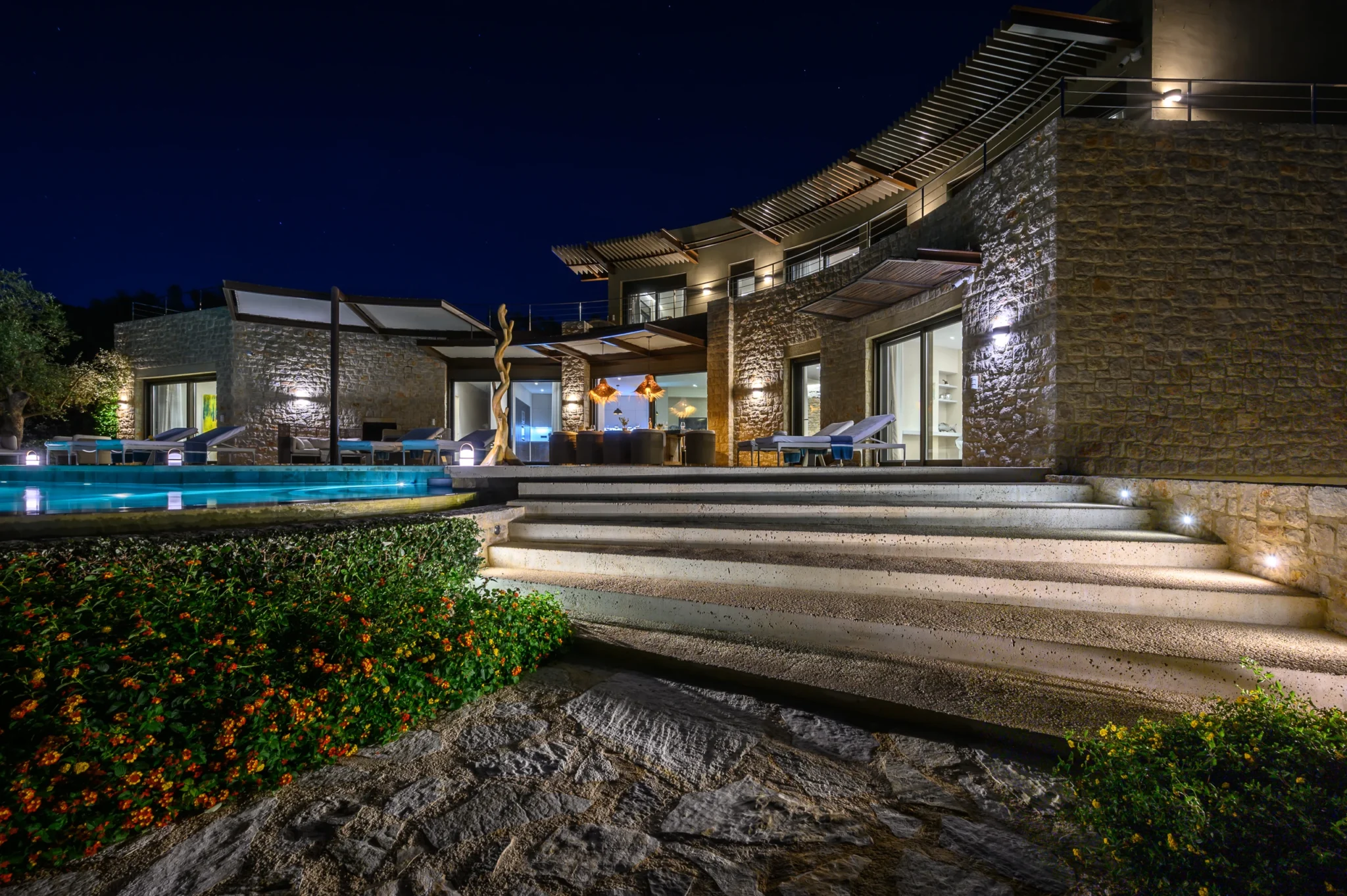 Illuminated stone facade with pergolas, broad curved steps and a covered dining terrace beside the swimming pool and planted garden at night at The Olive Retreat near Pylos.