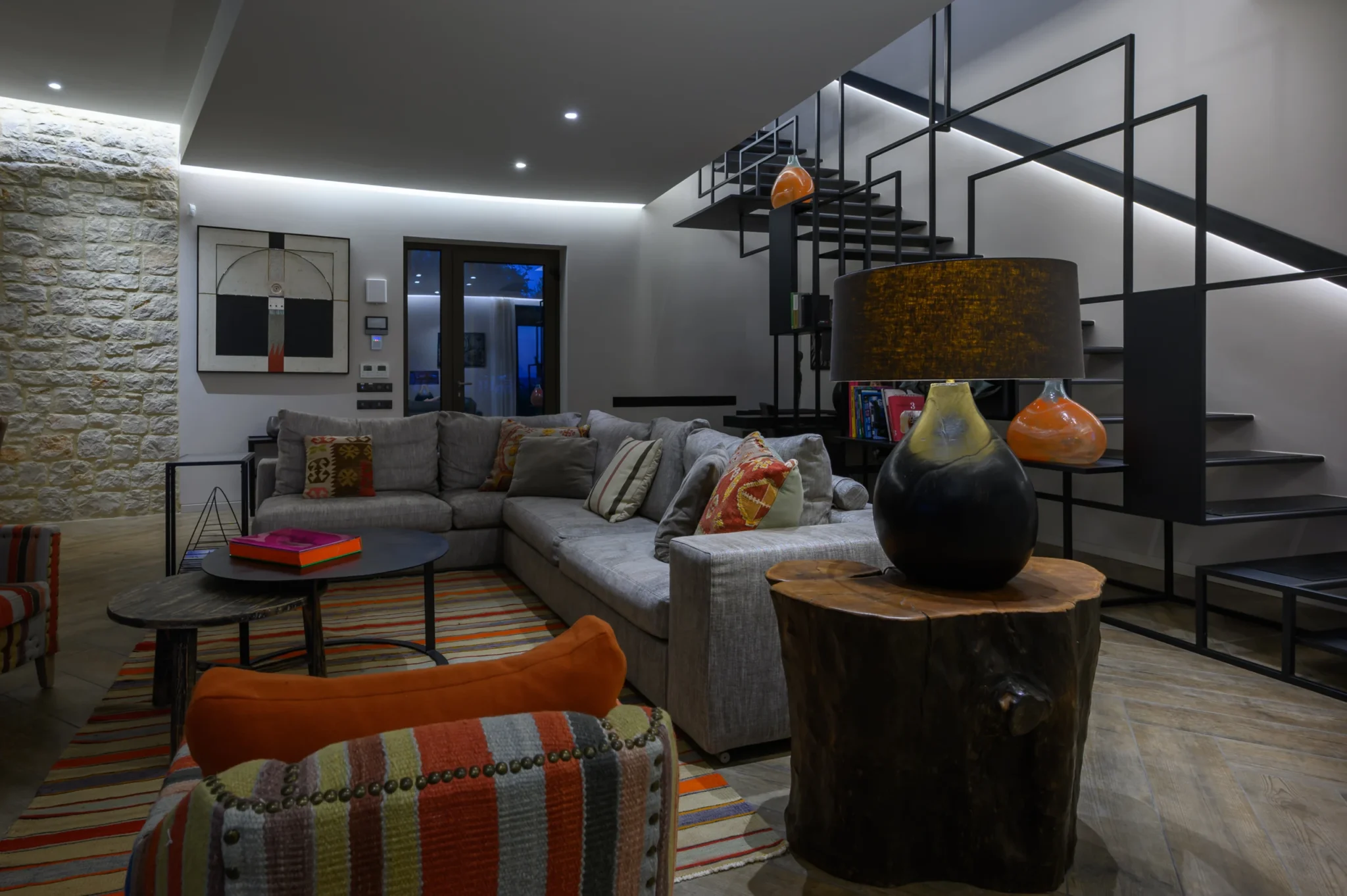 Grey corner sofa with patterned cushions and a striped rug beside a geometric steel staircase, with a ceramic lamp on a timber stump table at The Olive Retreat near Pylos.