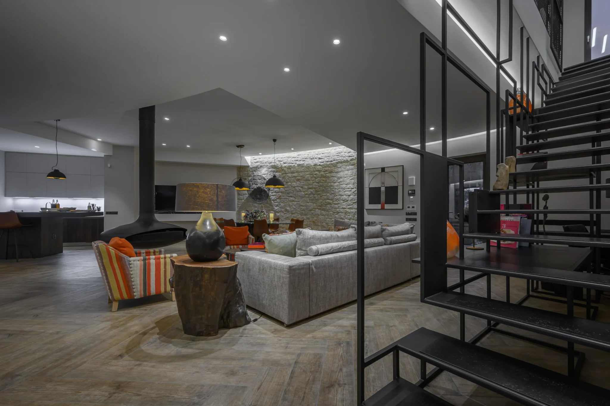Open-plan living room with grey seating, striped armchairs, a suspended black fireplace and a geometric steel staircase beside the kitchen and dining area at The Olive Retreat near Pylos.