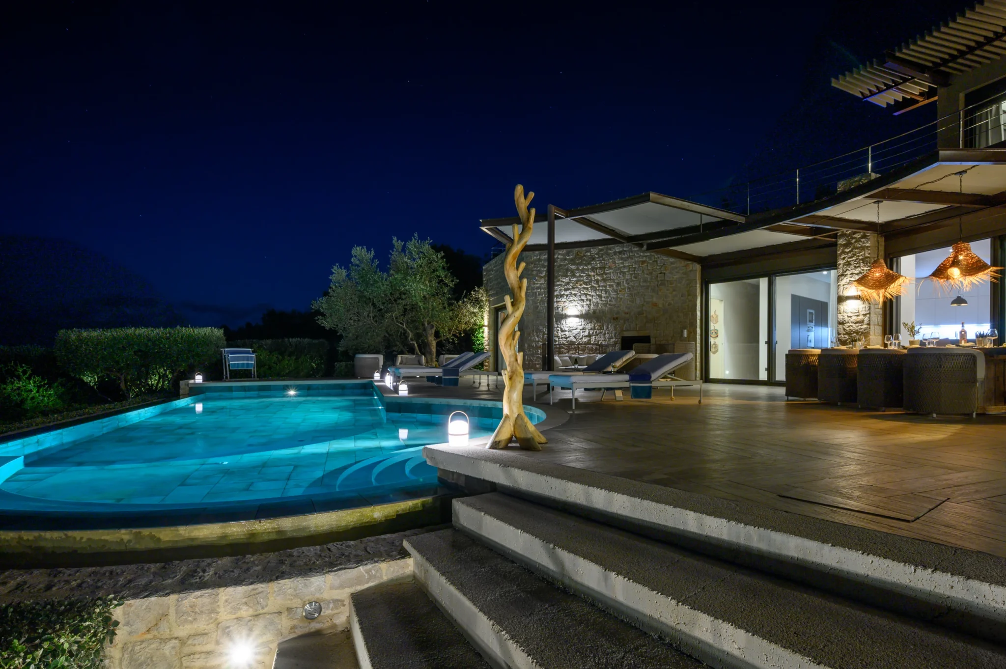 Illuminated swimming pool and stone facade beside a dining terrace with woven pendant lights, sun loungers and broad curved steps at night at The Olive Retreat near Pylos.