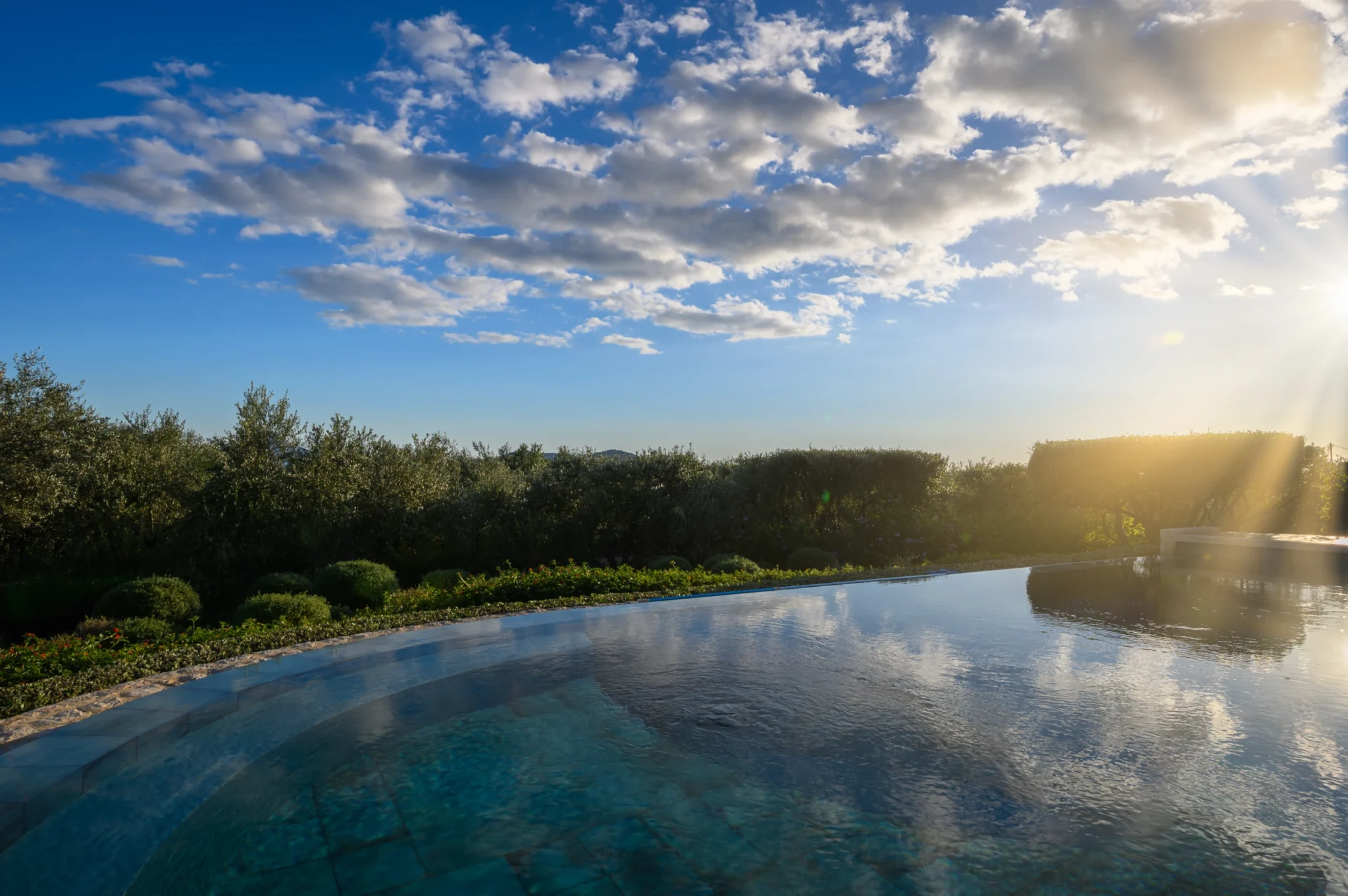 Swimming pool reflecting a cloud-filled sky, with olive trees and garden planting beyond in warm afternoon light at The Olive Retreat near Pylos.