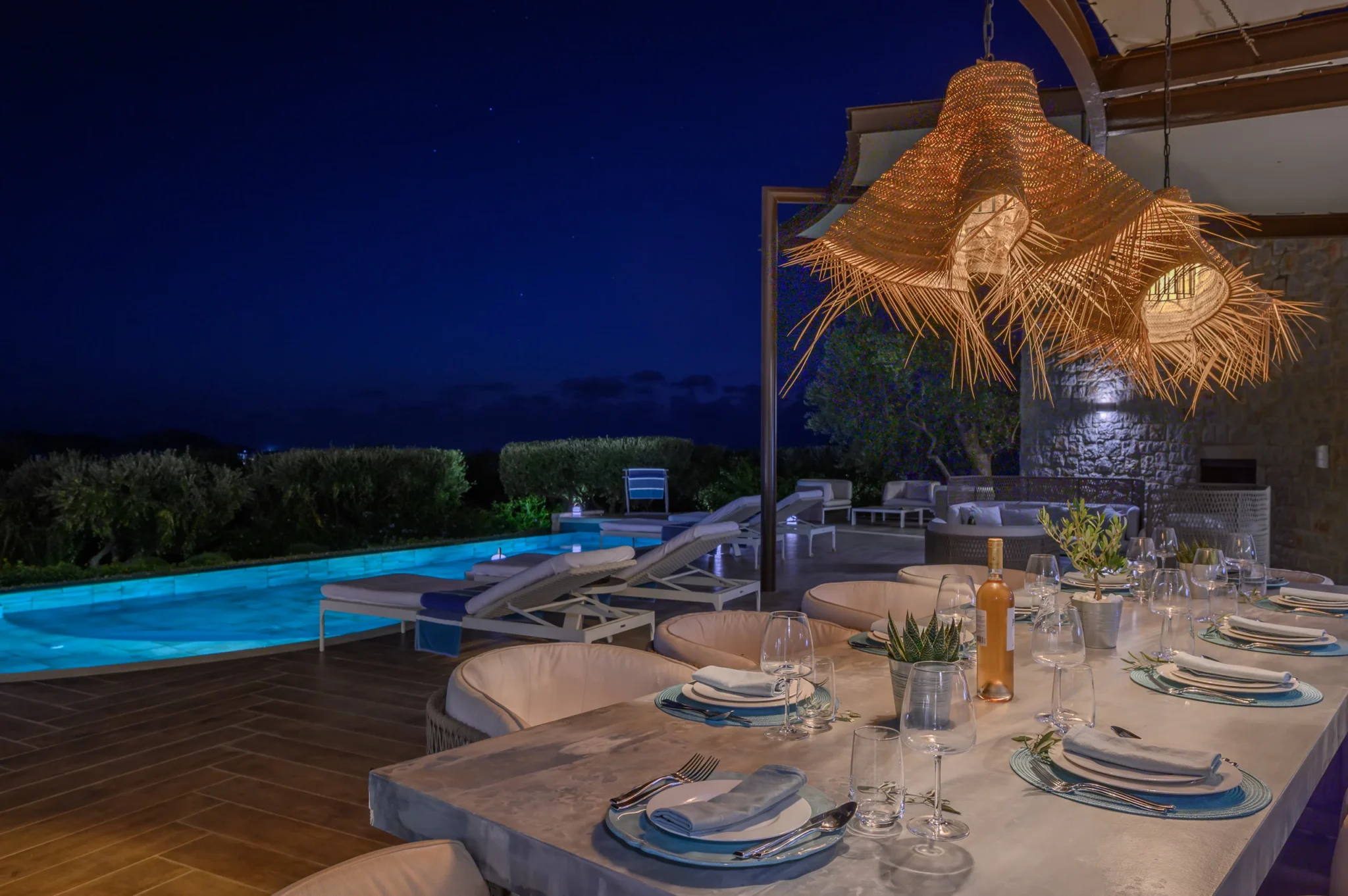 Outdoor dining table set with plates, glasses and blue placemats beneath glowing woven pendant lights, beside the illuminated pool at night at The Olive Retreat near Pylos.
