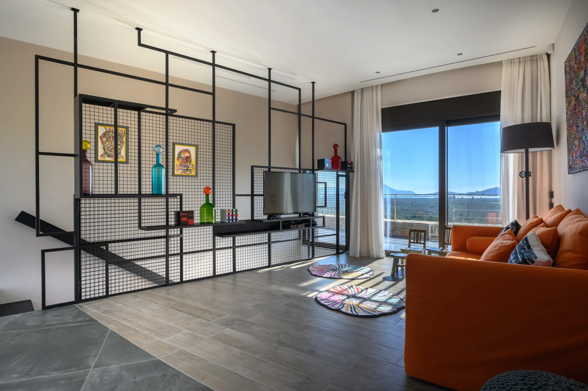 Sitting room with an orange sofa, geometric metal shelves displaying colourful glass bottles and artwork, and terrace doors overlooking the landscape at The Olive Retreat near Pylos.