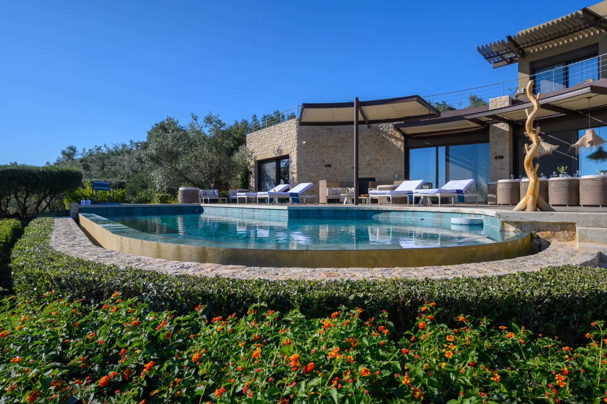 Stone retreat facade, shade canopies and upper terrace overlooking a curved swimming pool, with sun loungers and olive trees in daylight at The Olive Retreat near Pylos.