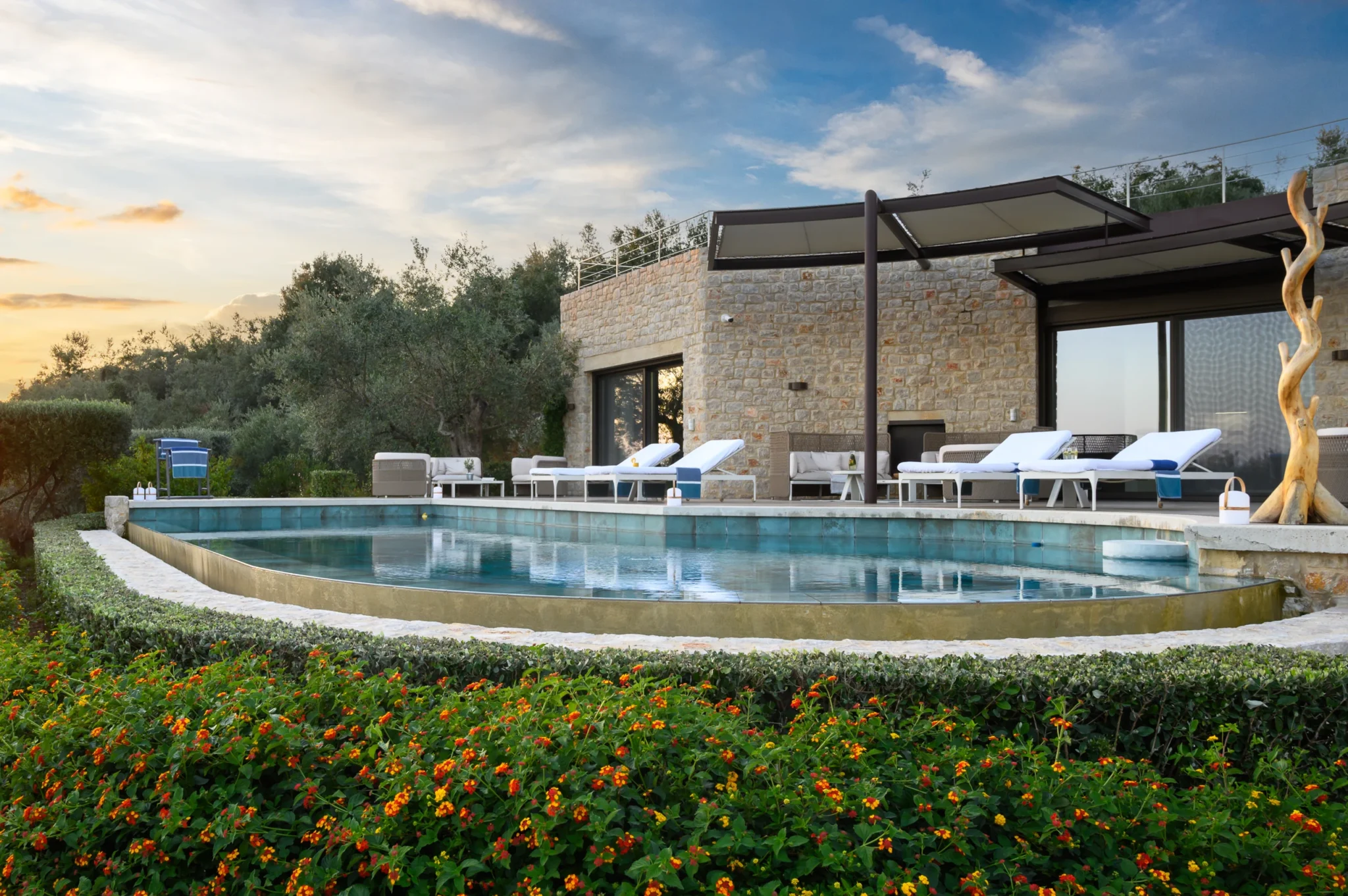 Stone facade and shaded terrace beside a curved swimming pool at dusk, with white sun loungers, olive trees and flowering garden planting at The Olive Retreat near Pylos.