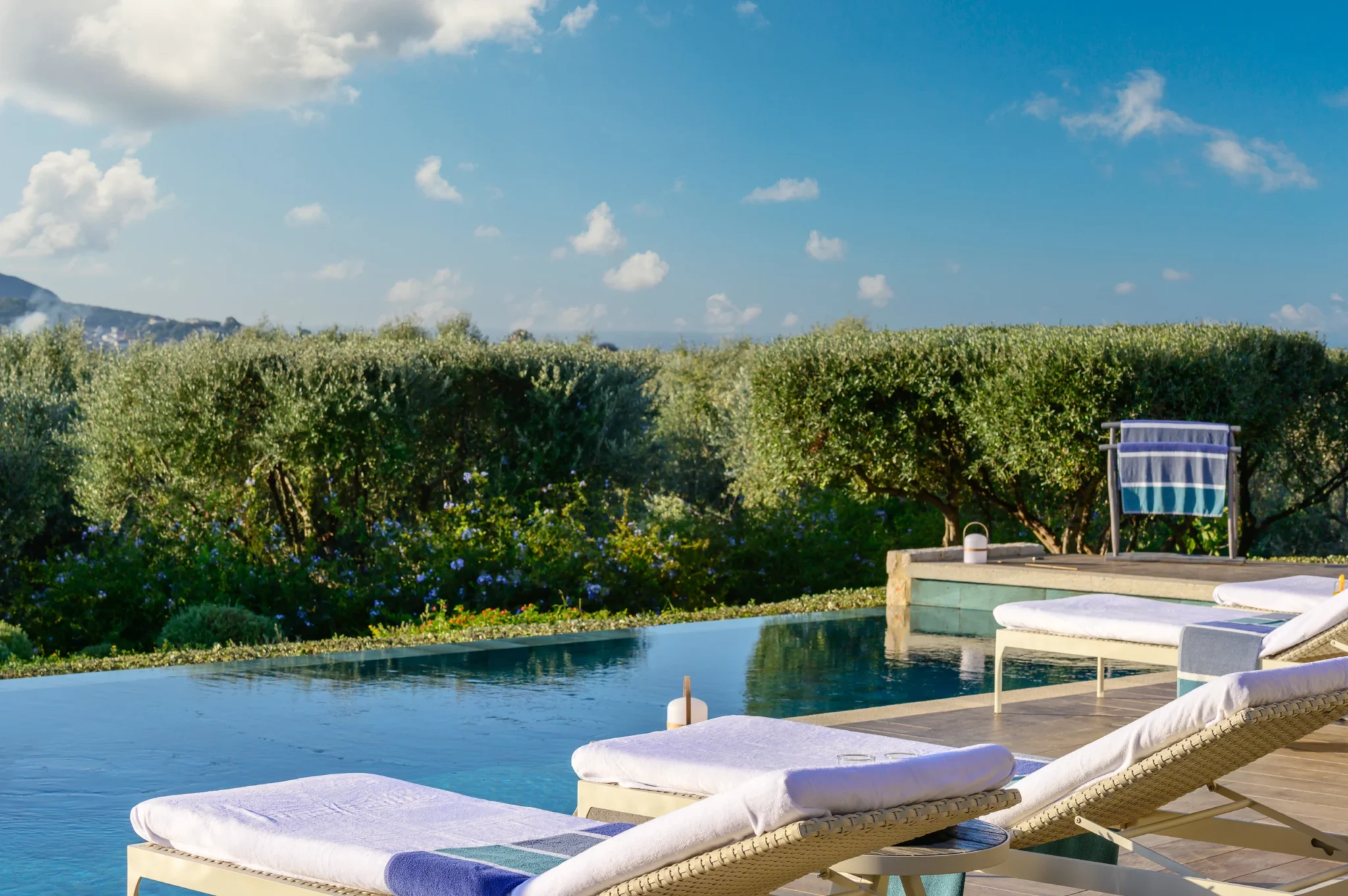 White cushioned sun loungers with blue towels beside the swimming pool, with olive trees and flowering garden planting beyond at The Olive Retreat near Pylos.