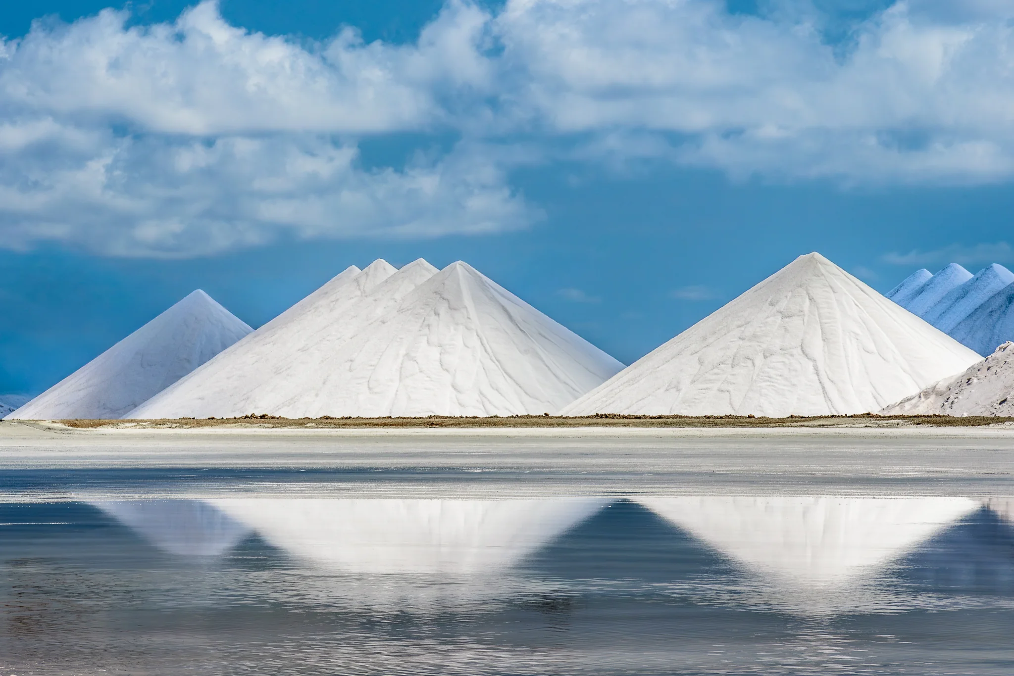 White salt mounds reflected in shallow water beneath scattered clouds