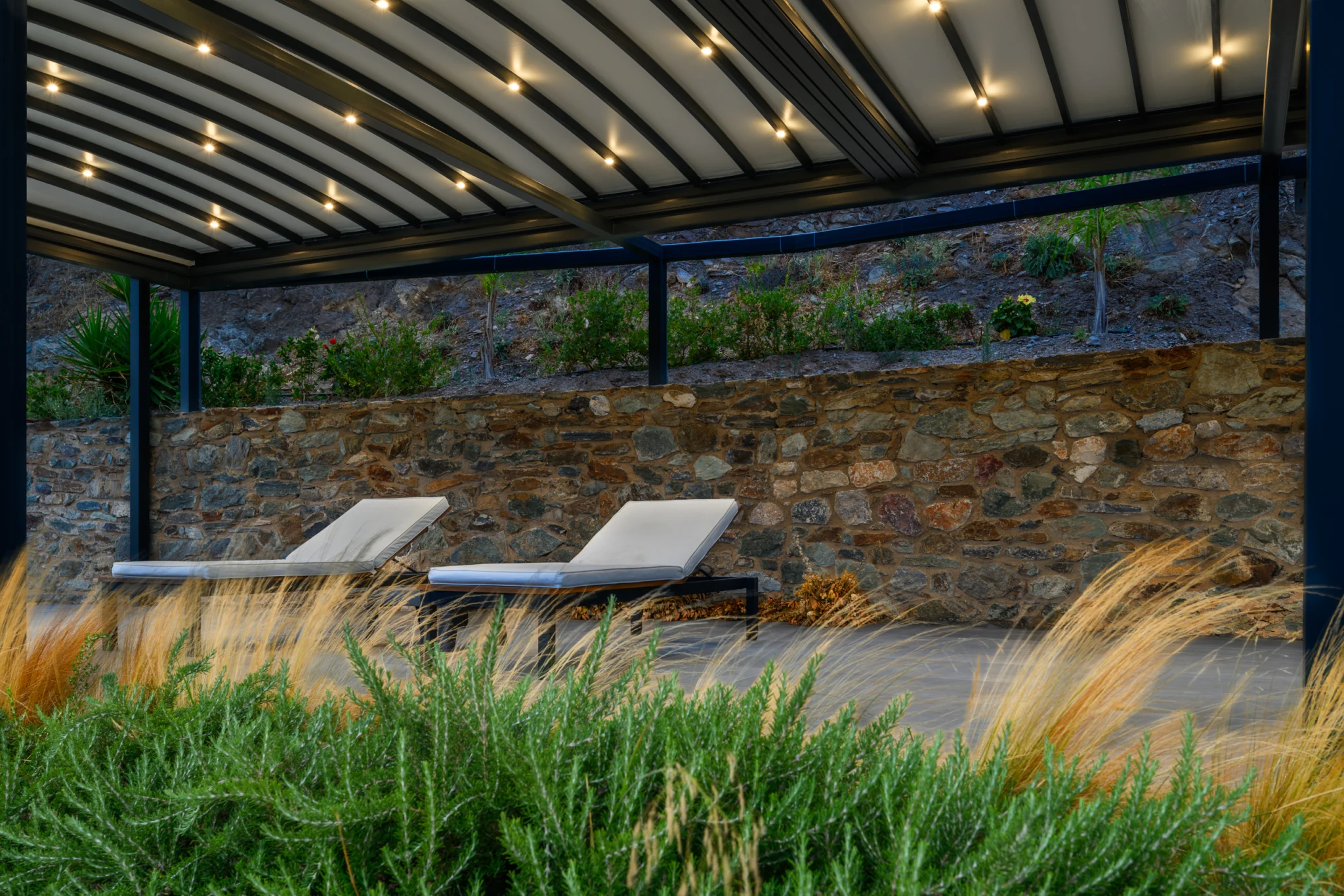 Illuminated pergola with loungers, ornamental grasses and a stone wall at Villa in Athens, Athens, Attica, Greece.