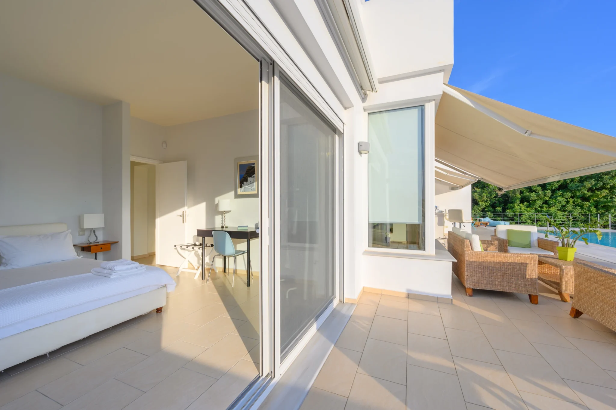 White bedroom with folded towels and a desk beside sliding glass doors opening to wicker terrace seating beneath a cream awning at Villa Bel Air in Porto Heli.