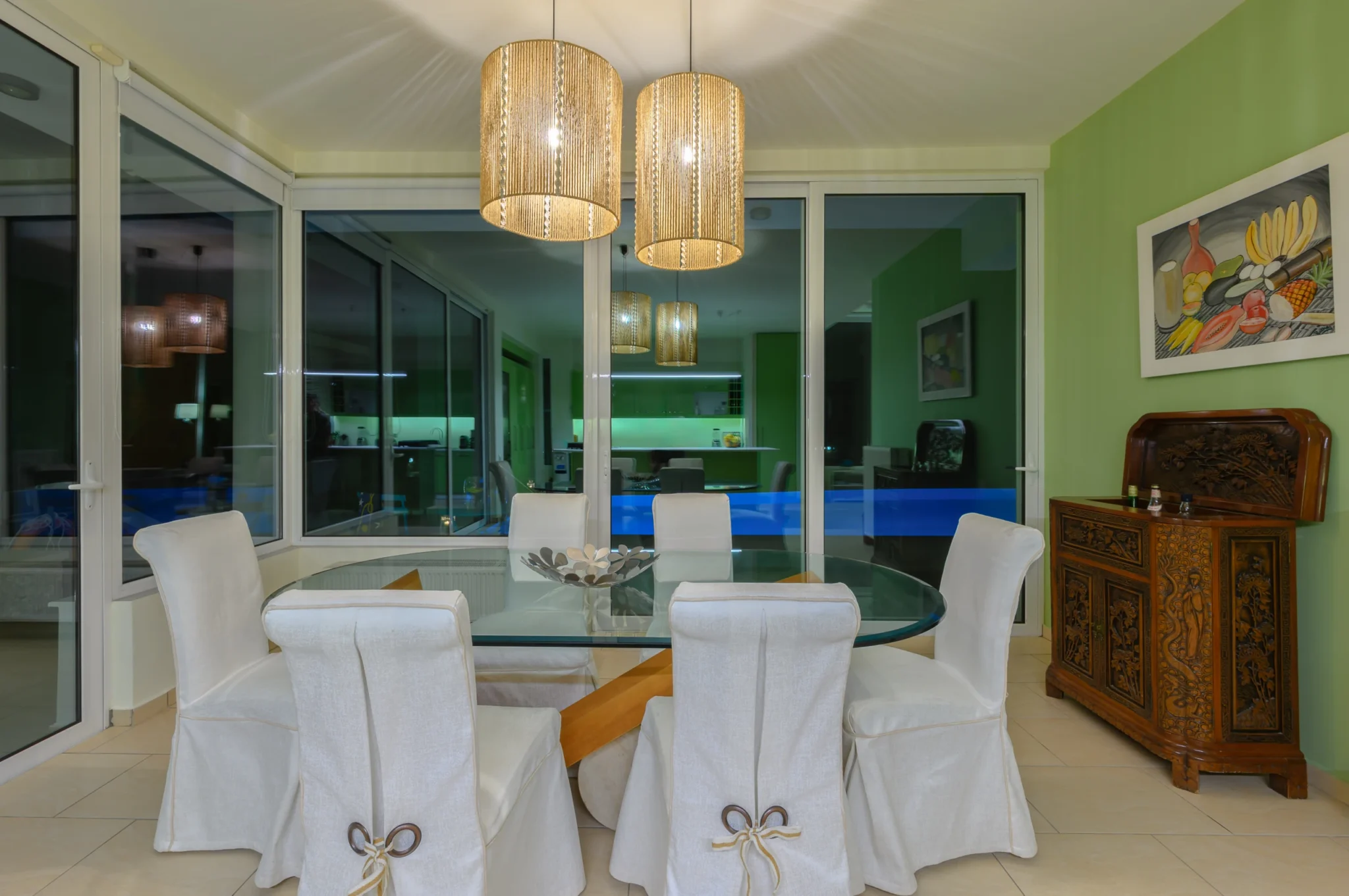 Glass dining table with white covered chairs beneath two glowing woven pendants, beside a green wall and carved timber cabinet in the evening at Villa Bel Air in Porto Heli.