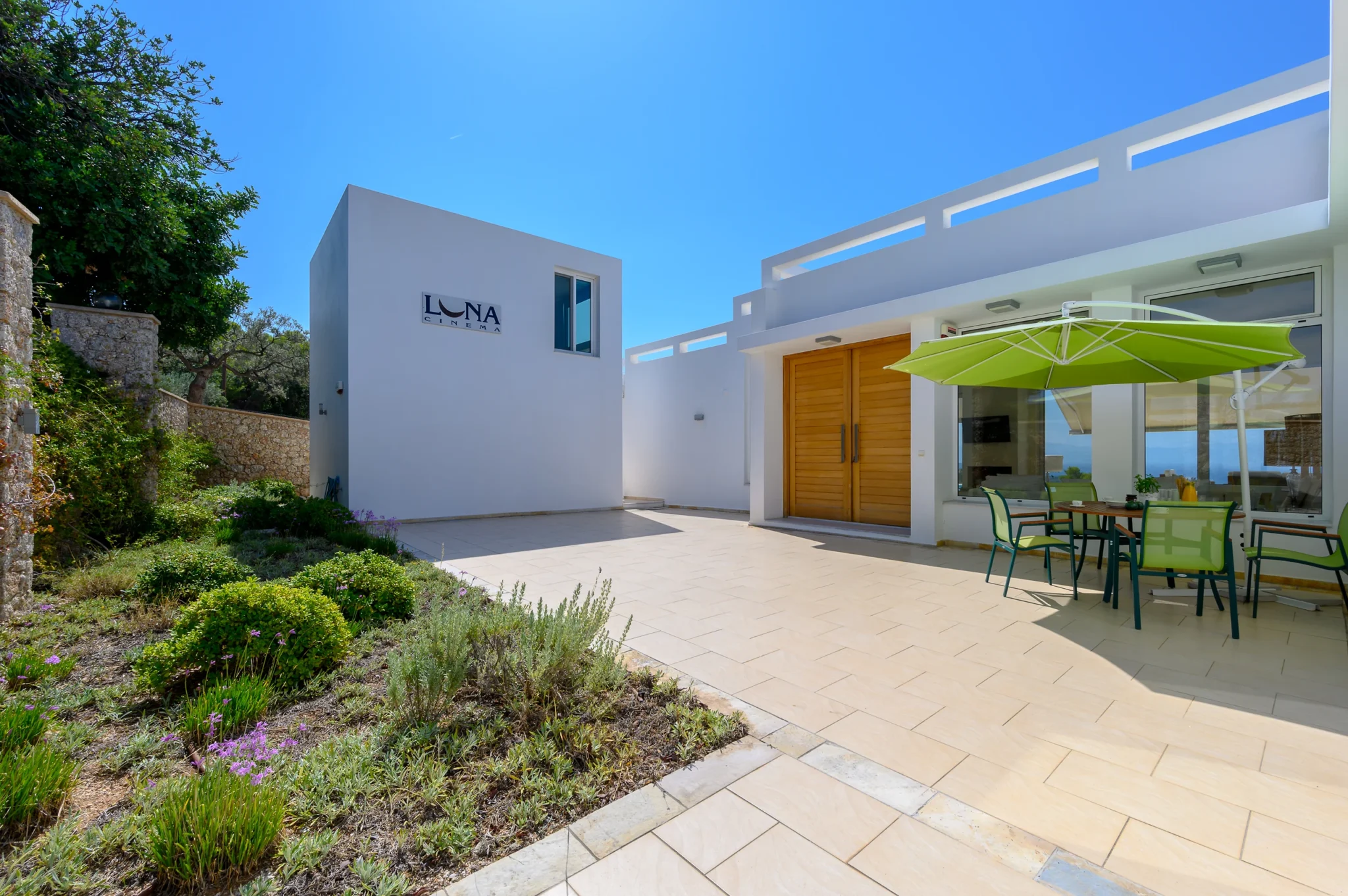 White entrance courtyard with timber double doors, planted borders and a dining table beneath a lime-green parasol at Villa Bel Air in Porto Heli.