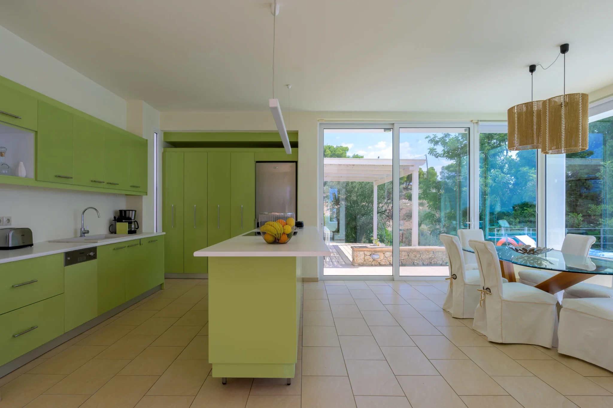 Lime-green kitchen with a white-topped island and fruit bowl beside a glass dining table, white chairs and large garden-facing doors at Villa Bel Air in Porto Heli.