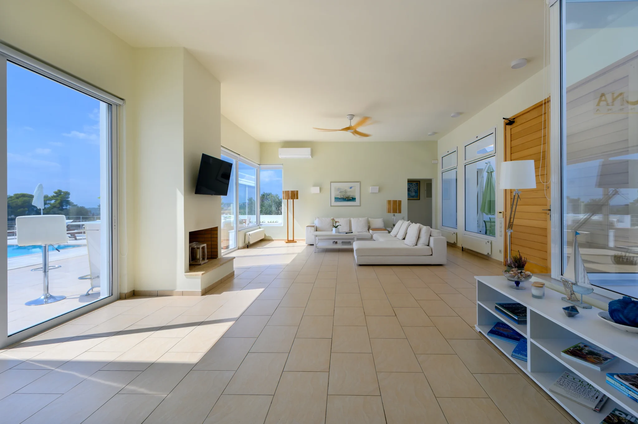 Long living room with a white corner sofa, fireplace, timber entrance doors and broad glass doors overlooking the pool terrace at Villa Bel Air in Porto Heli.