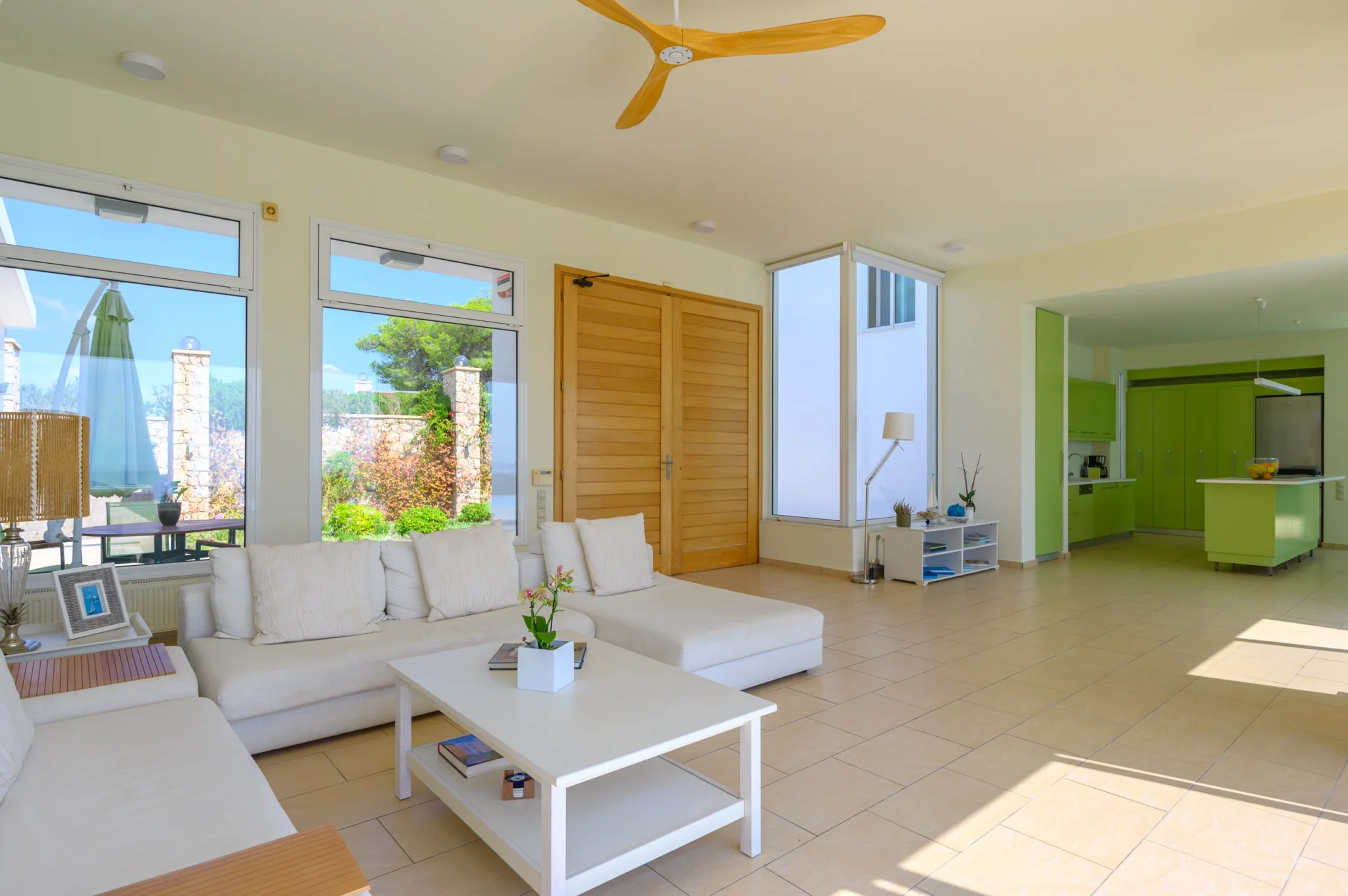 White corner sofa beside timber double doors in an open living area, with a lime-green kitchen beyond and garden views through tall windows at Villa Bel Air in Porto Heli.
