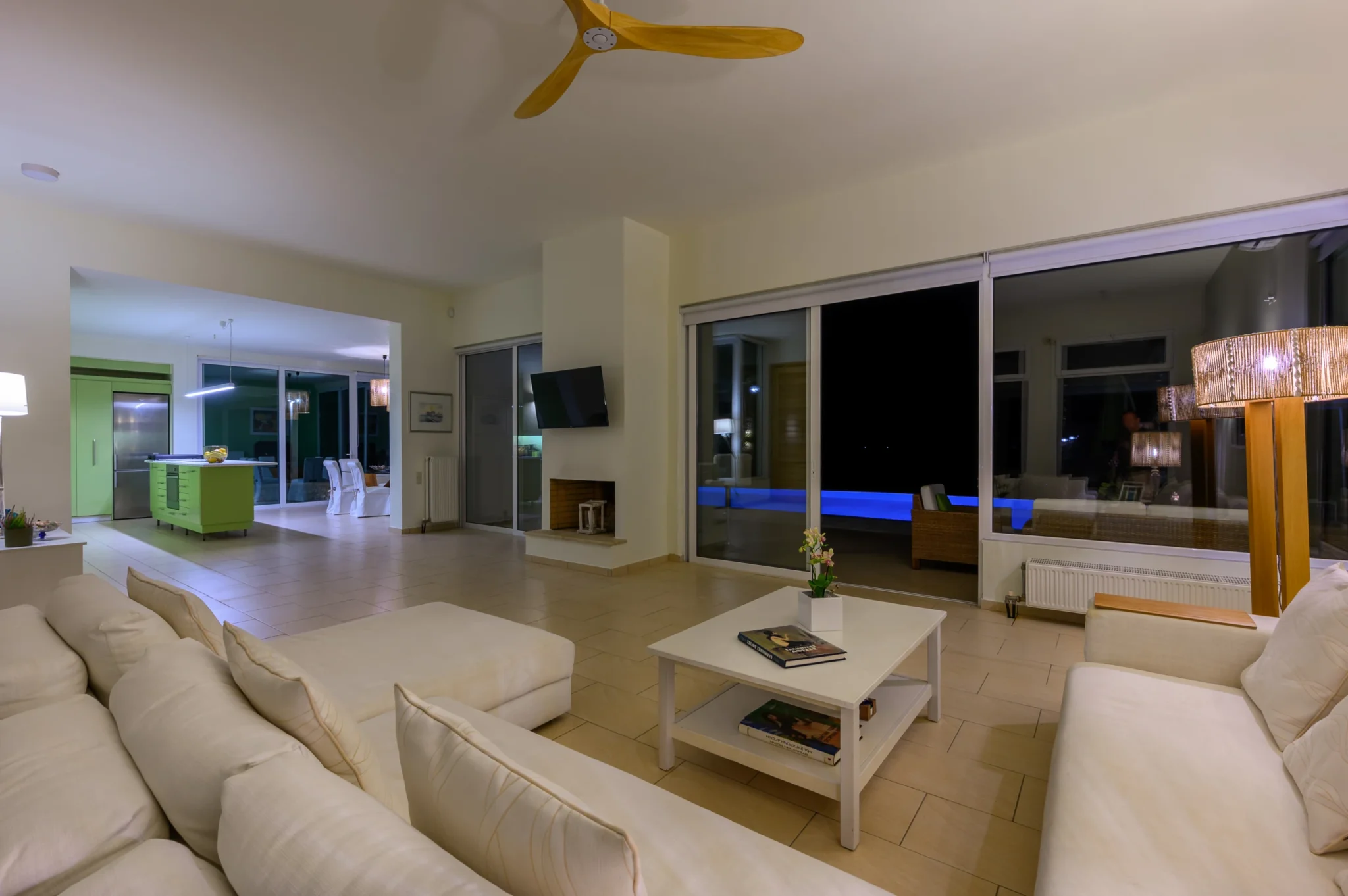 White living-room seating, a fireplace and woven floor lamp beside glass doors revealing the blue-lit pool, with the green kitchen beyond at Villa Bel Air in Porto Heli.