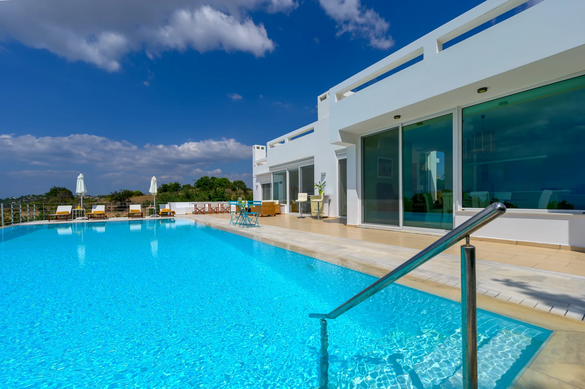 Turquoise swimming pool with a stainless-steel handrail beside the white facade, broad glass doors and a terrace with sun loungers at Villa Bel Air in Porto Heli.