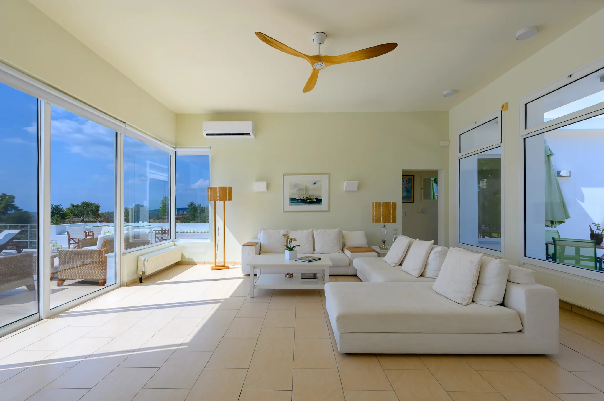White sofas and a coffee table in a bright living room, with a wooden ceiling fan and sunlight entering through broad terrace windows at Villa Bel Air in Porto Heli.