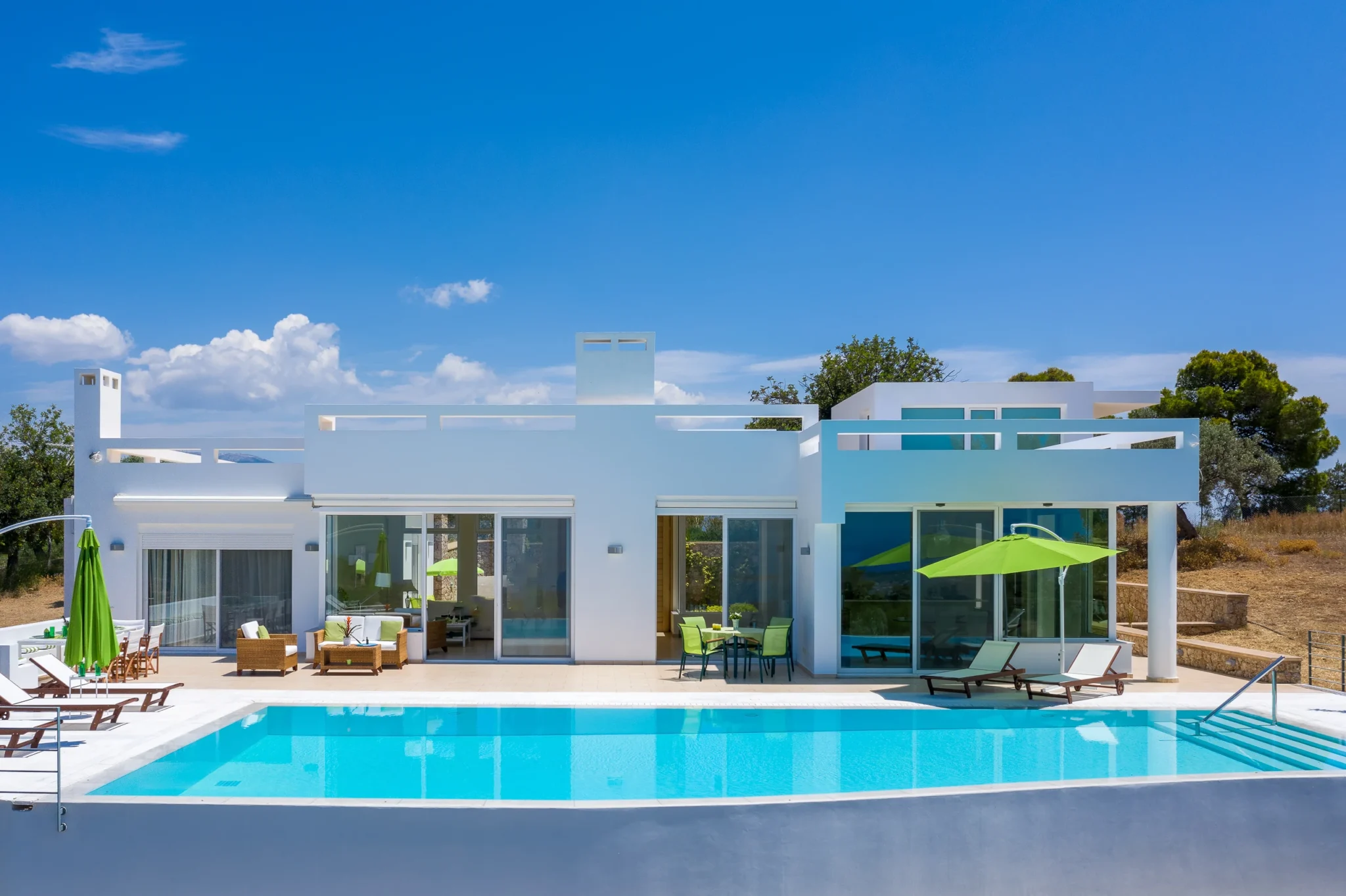 White villa with broad glazed openings, lime-green parasols and sun loungers overlooking a turquoise swimming pool at Villa Bel Air in Porto Heli.