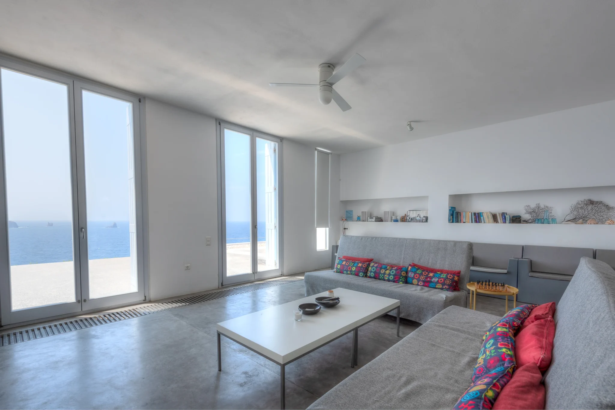 Grey sofas with colourful cushions and recessed bookshelves in a white lounge overlooking the sea at Villa Edge in Paros.