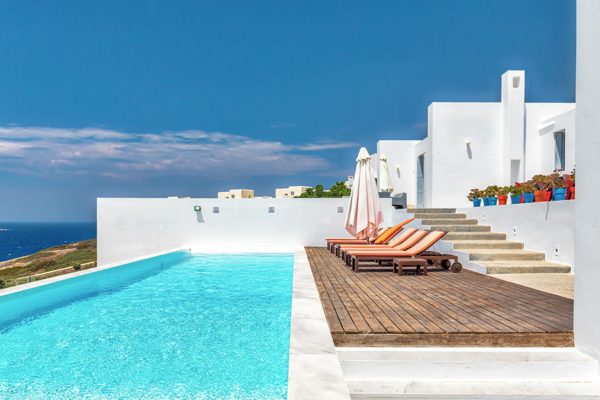 Turquoise swimming pool beside a timber deck with orange loungers, white umbrellas and stepped terraces at Villa Edge in Paros.