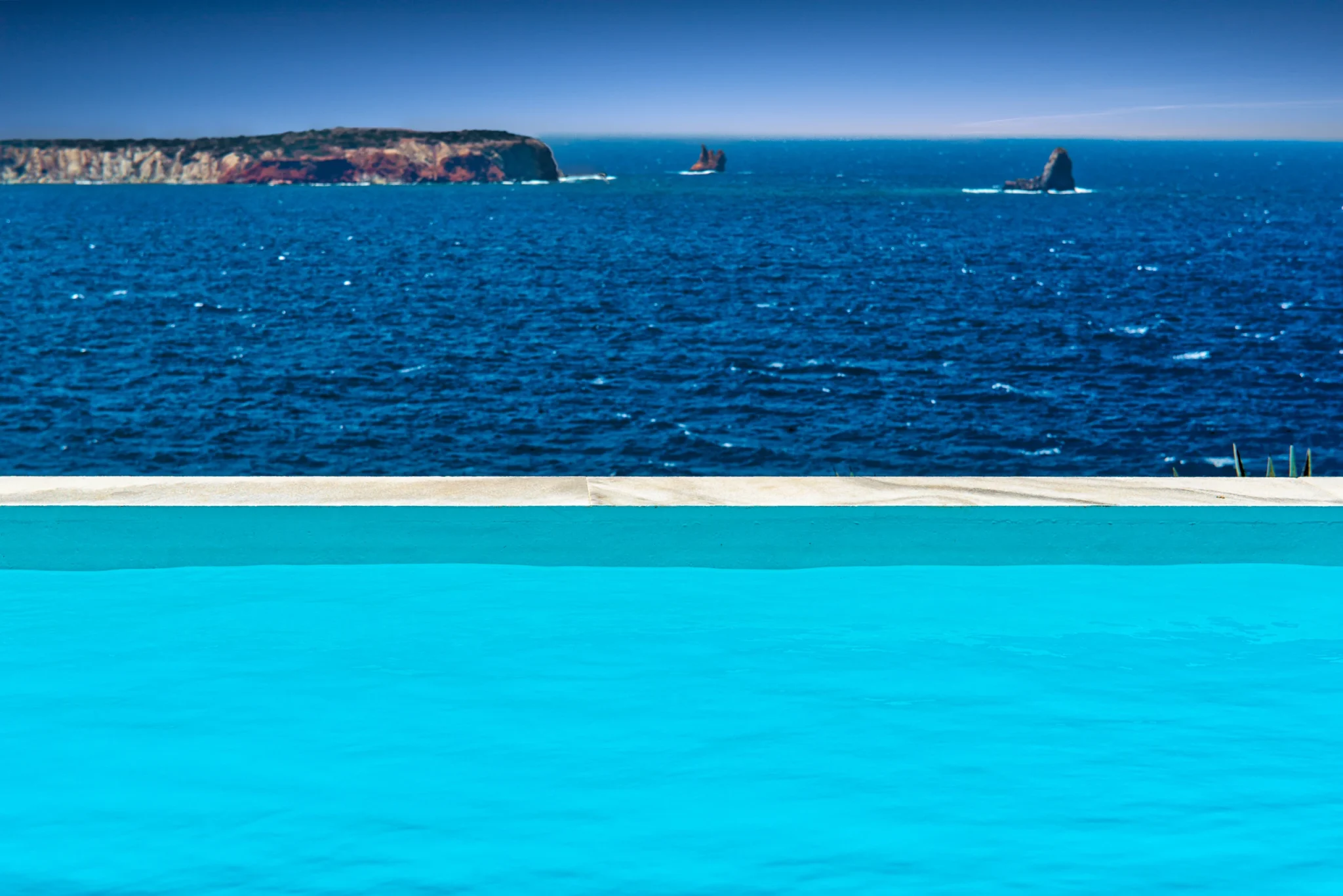 Turquoise pool water and a pale pool edge frame the deep blue sea and rocky islets beyond Villa Edge in Paros.