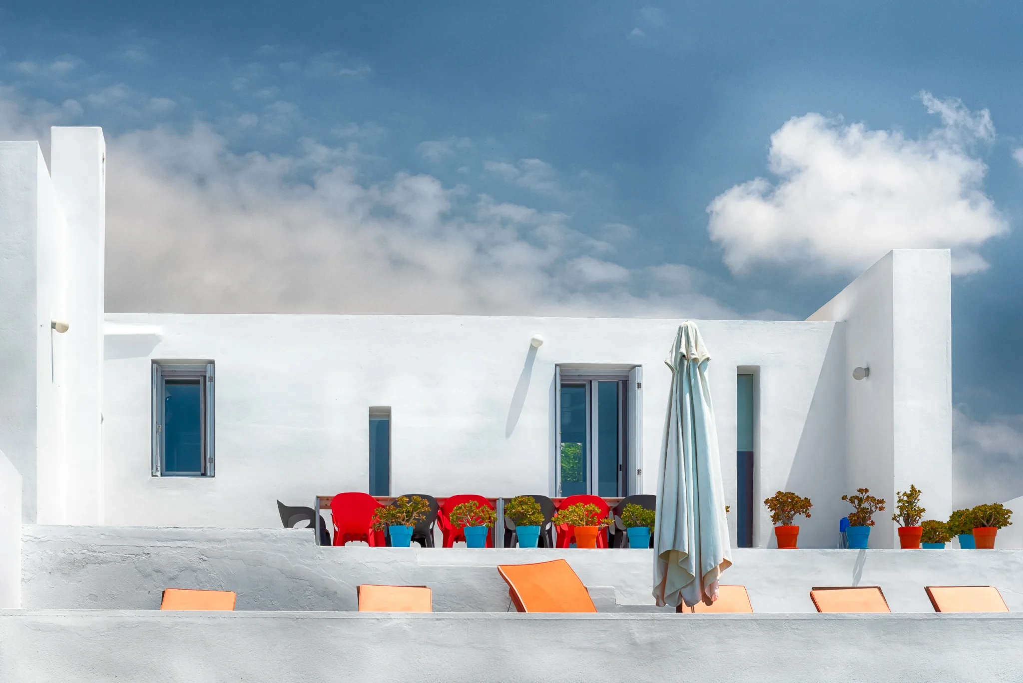 White villa facade with colourful dining chairs and plant pots above orange poolside loungers at Villa Edge in Paros.