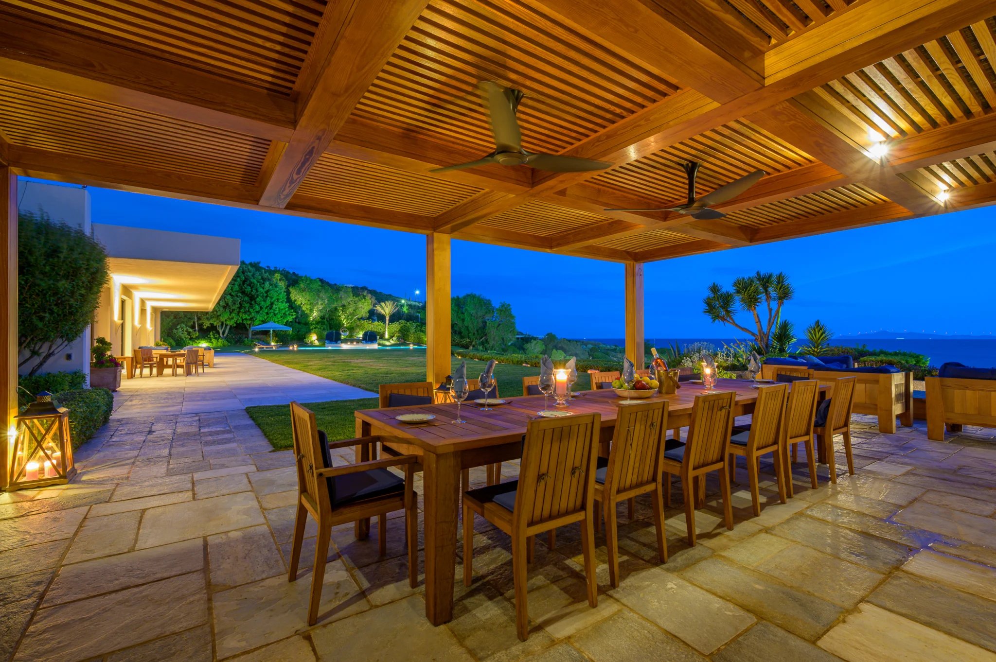 Outdoor dining under a lit timber pergola beside the lawn at twilight at Villa Icarus.