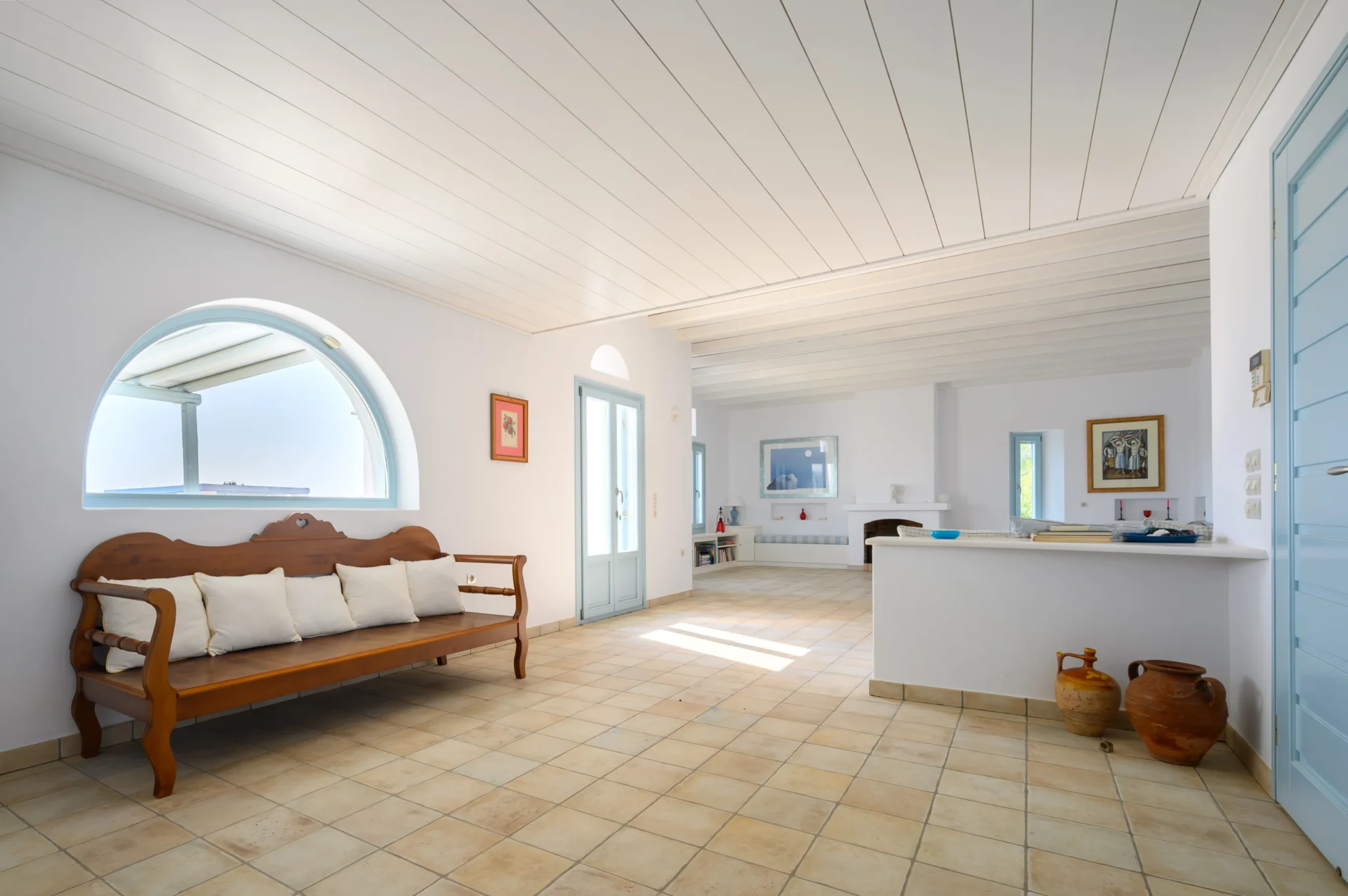 Wooden bench with white cushions beneath a pale blue arched window in the bright interior of Villa Lumina in Paros.