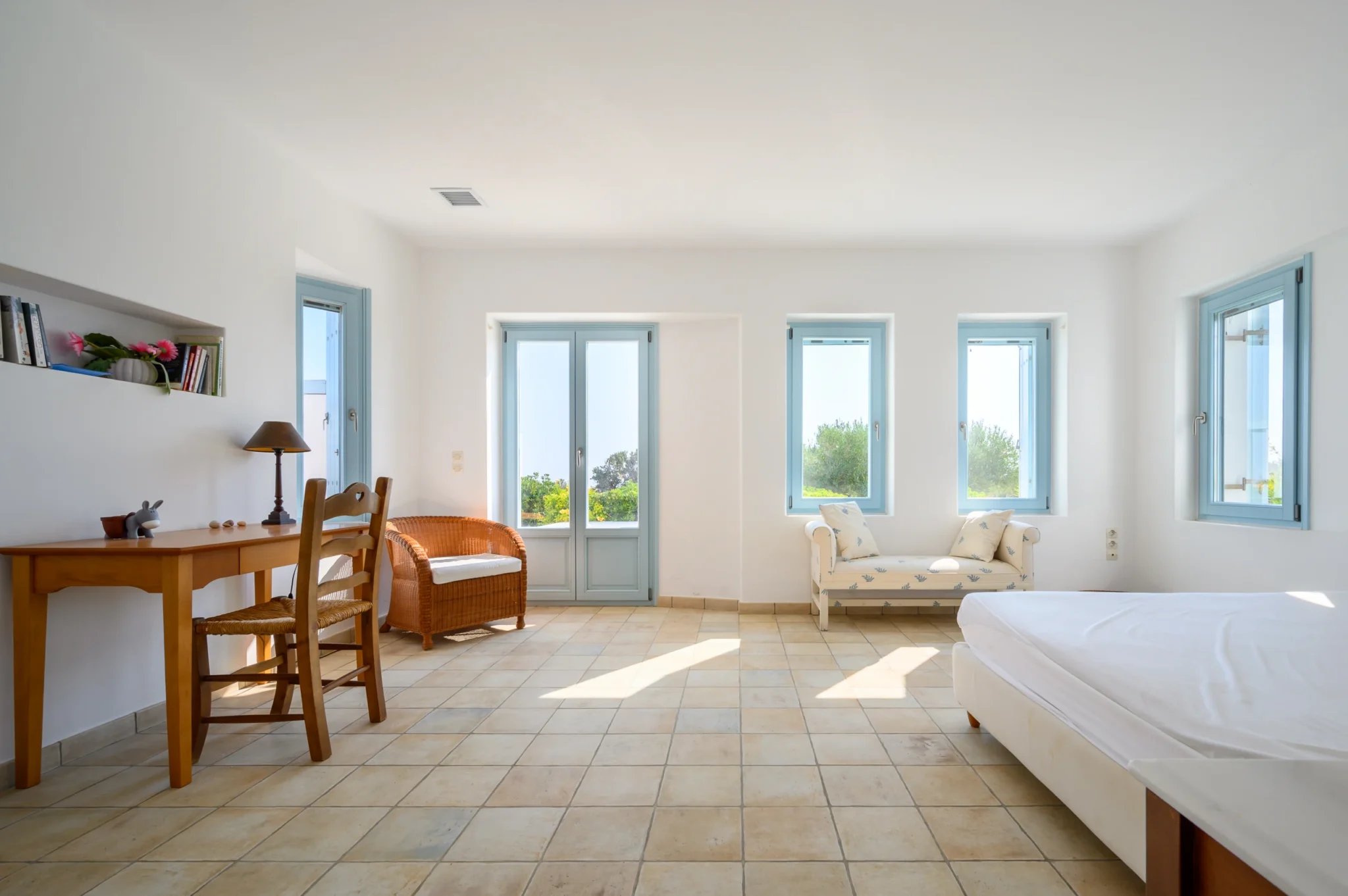 Bright white bedroom with pale blue windows and doors, a wooden desk, wicker chair and upholstered settee at Villa Lumina in Paros.