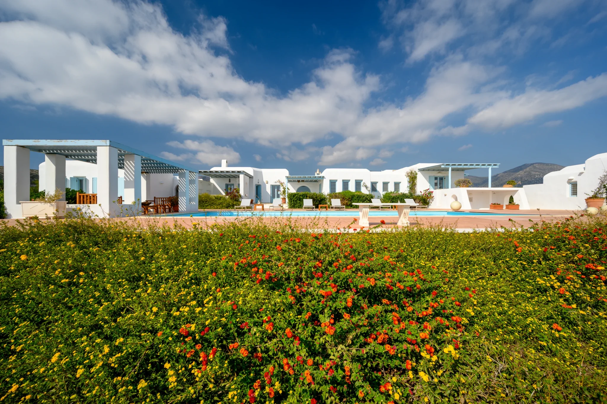Wide view of Villa Lumina in Paros, with white buildings, pale blue pergolas, a swimming pool and red and yellow garden flowers.