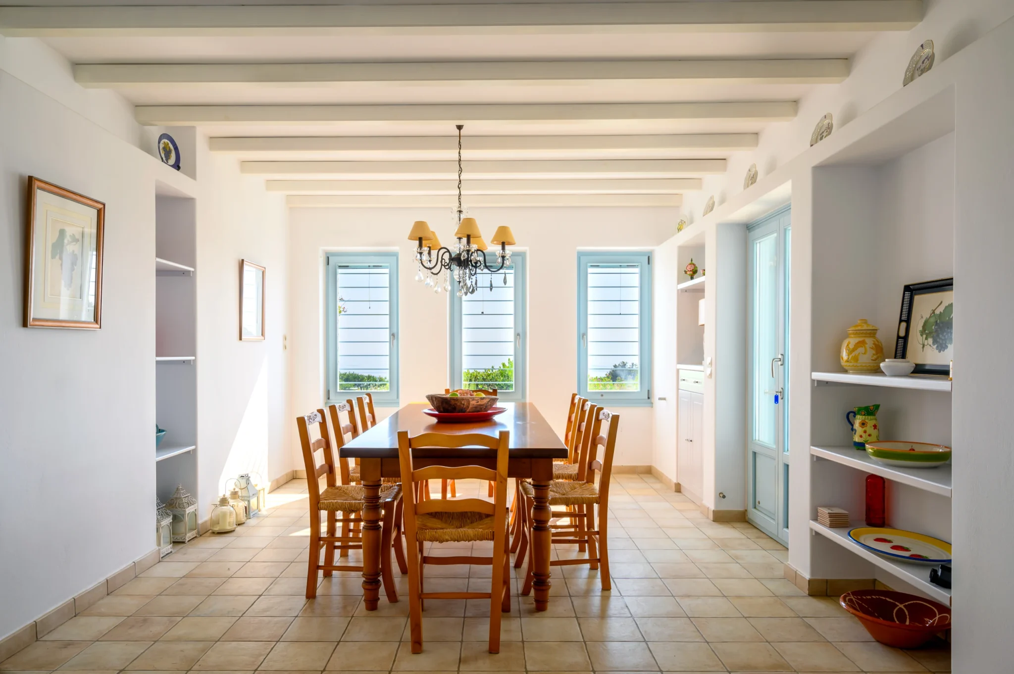 Wooden dining table and chairs beneath white ceiling beams and a chandelier, with pale blue windows at Villa Lumina in Paros.