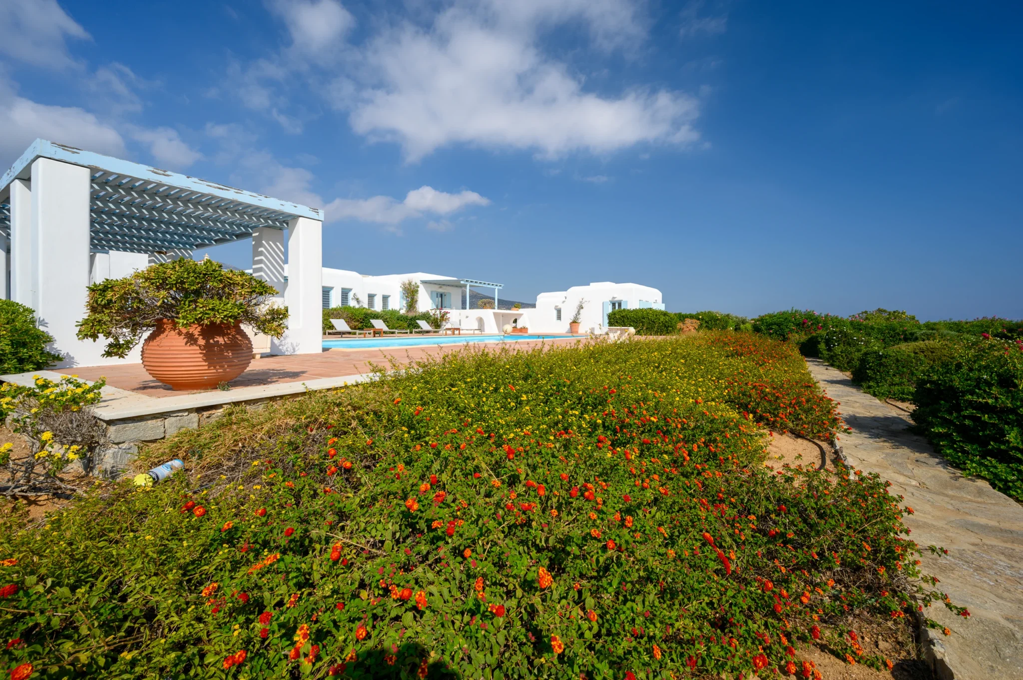 Stone garden path beside red and yellow flowers, a large terracotta planter and a poolside pergola at Villa Lumina in Paros.