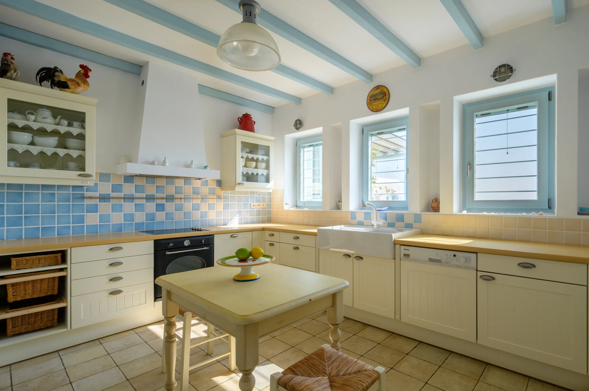 Cream kitchen cabinetry, timber worktops, a farmhouse sink and blue tiled details beneath pale blue ceiling beams at Villa Lumina in Paros.