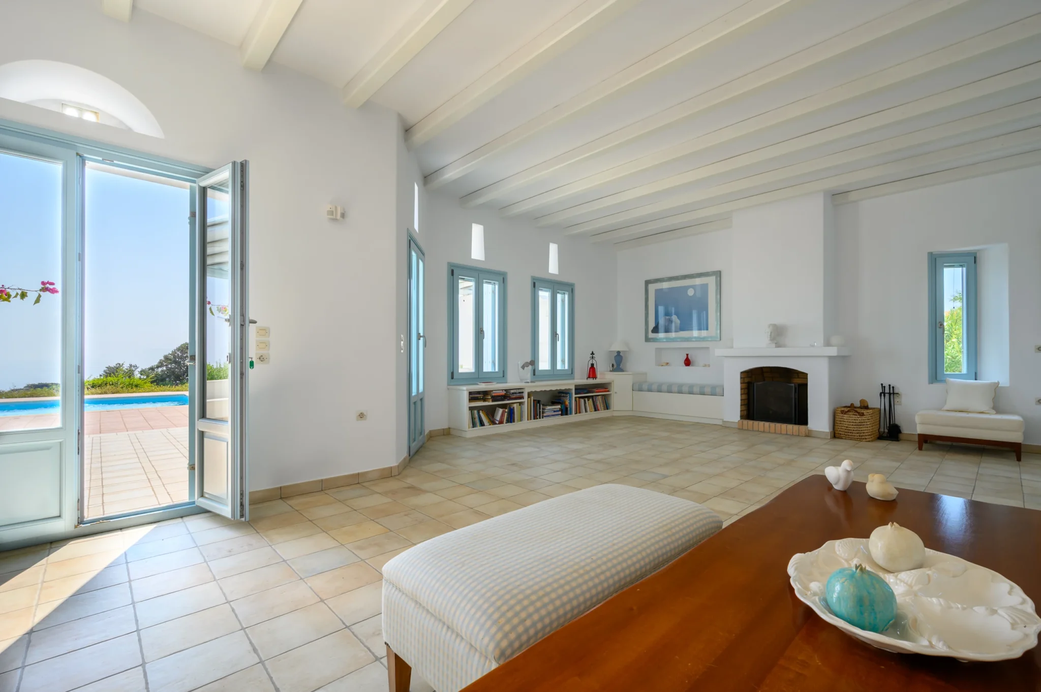White living room with exposed ceiling beams, pale blue doors, a fireplace and an open doorway to the pool terrace at Villa Lumina in Paros.