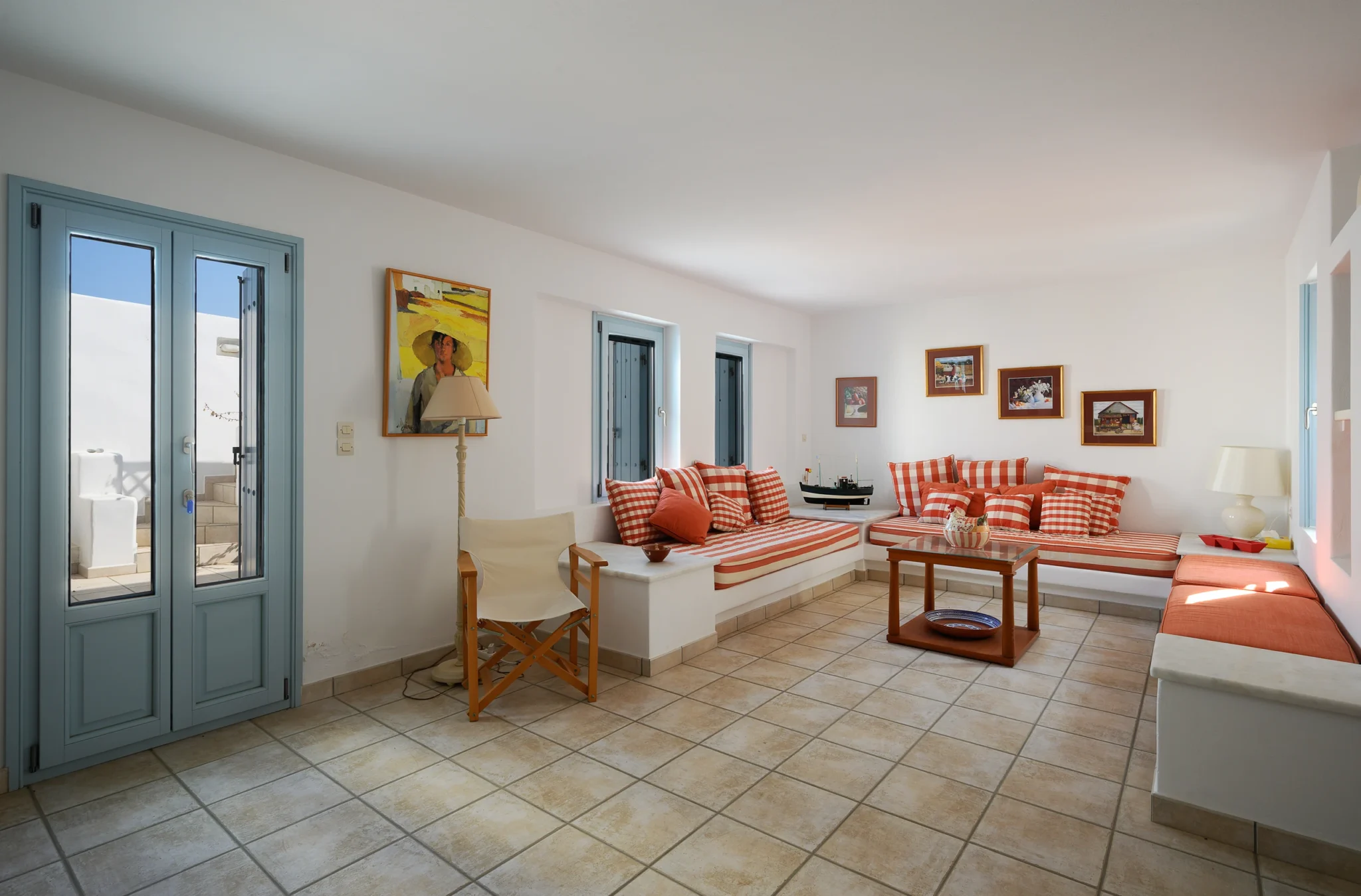 White built-in lounge seating with red and white cushions, a wooden coffee table and pale blue doors at Villa Lumina in Paros.