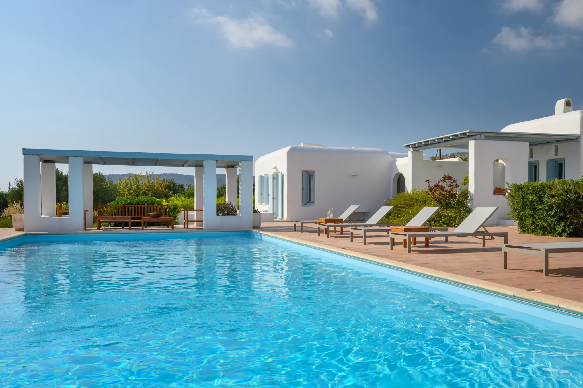 Swimming pool beside a terracotta tiled terrace, sun loungers and a pale blue pergola with a wooden bench at Villa Lumina in Paros.
