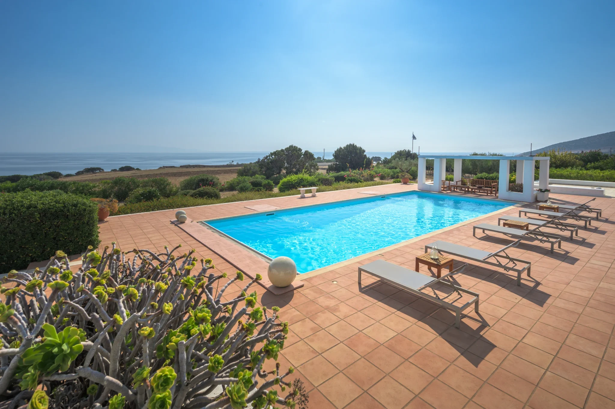 Elevated view across the swimming pool, loungers and pergola towards the sea and surrounding landscape at Villa Lumina in Paros.