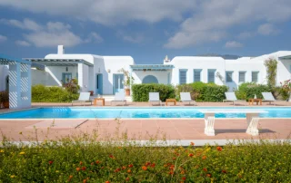 White Cycladic villa with pale blue windows and pergolas beside a turquoise swimming pool and flowering garden at Villa Lumina in Paros.