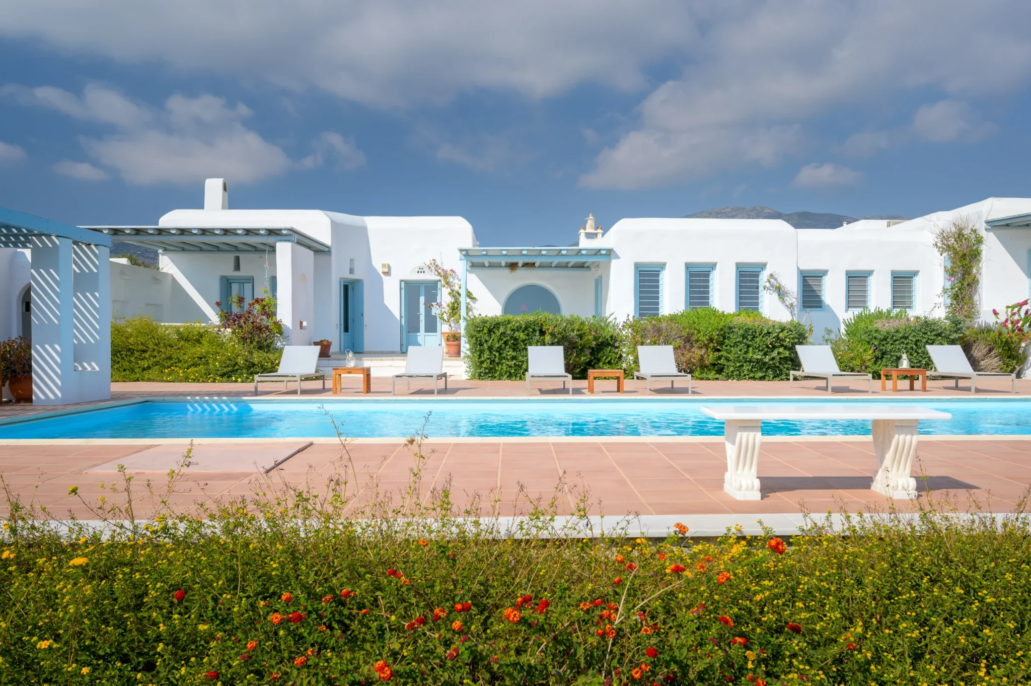 White Cycladic villa with pale blue windows and pergolas beside a turquoise swimming pool and flowering garden at Villa Lumina in Paros.
