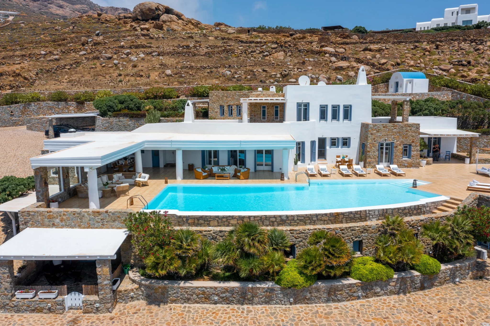 Aerial view of the villa facade, swimming pool, covered terraces and planted stone retaining walls against the rocky hillside at Villa Mustique in Mykonos.