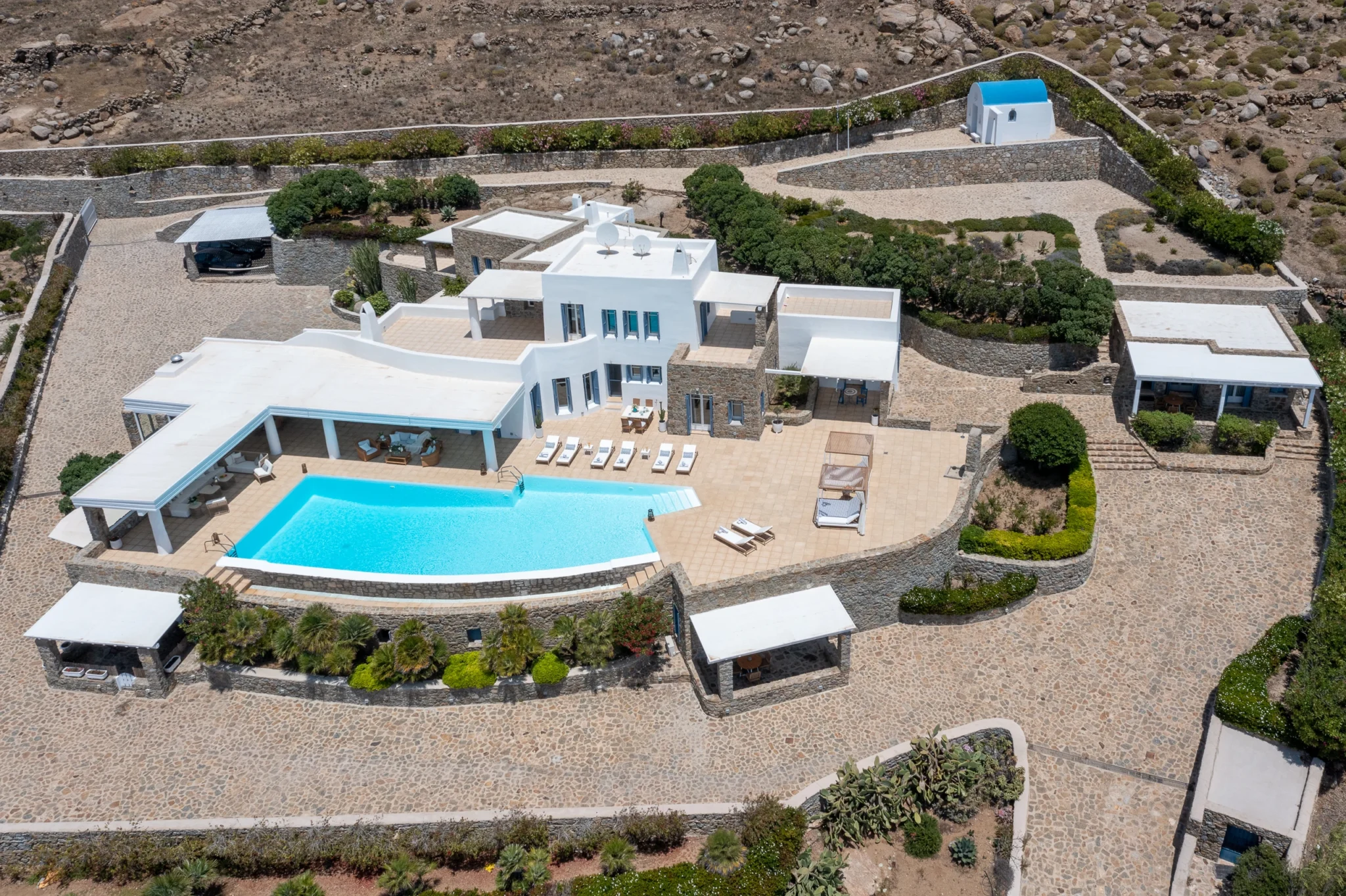 Aerial view of the white and stone villa, swimming pool, terraces, planted gardens and surrounding stone paths at Villa Mustique in Mykonos.