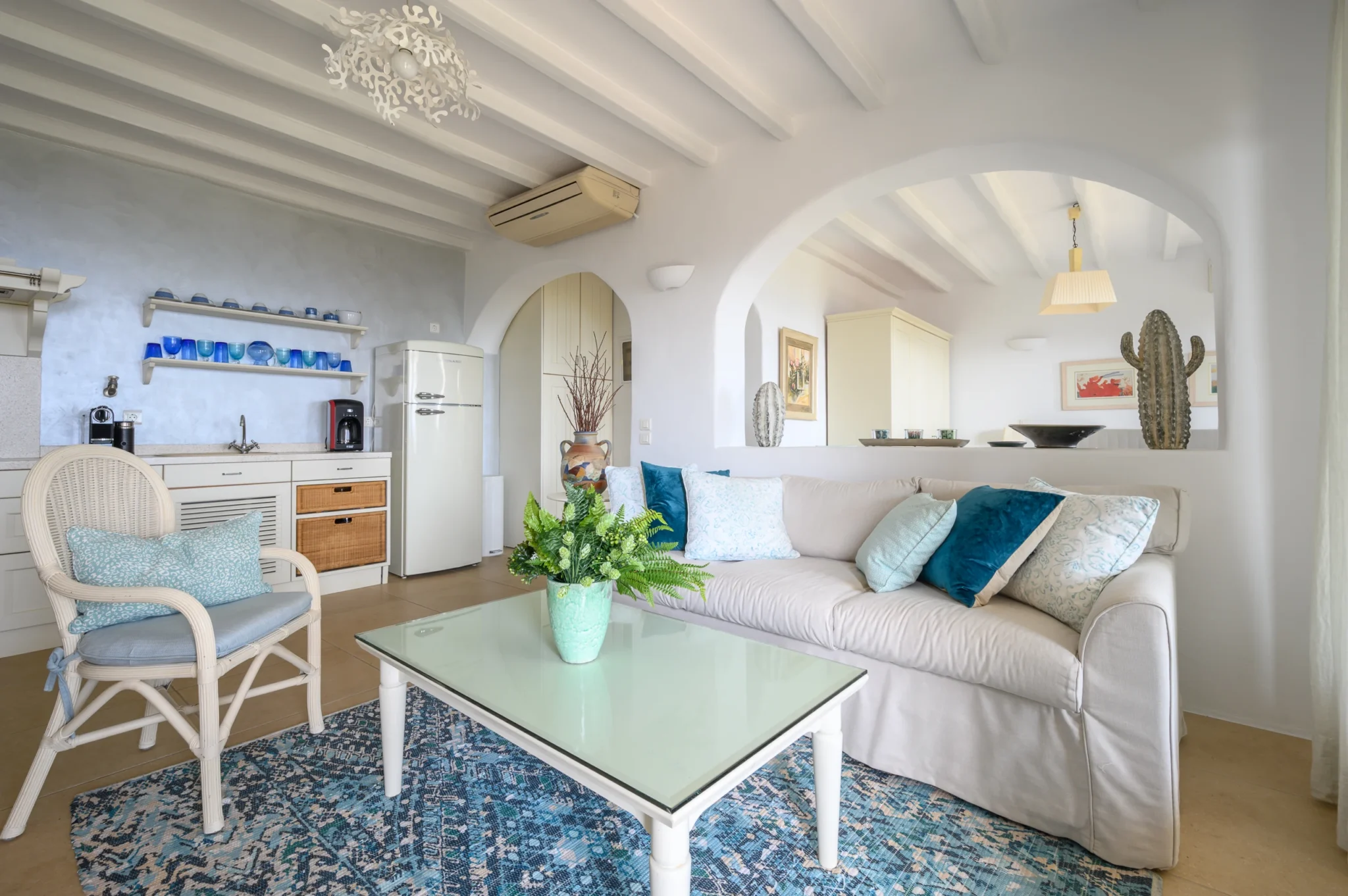 White living area with turquoise cushions and a blue patterned rug, beside a kitchenette and arched opening to the bedroom at Villa Mustique in Mykonos.