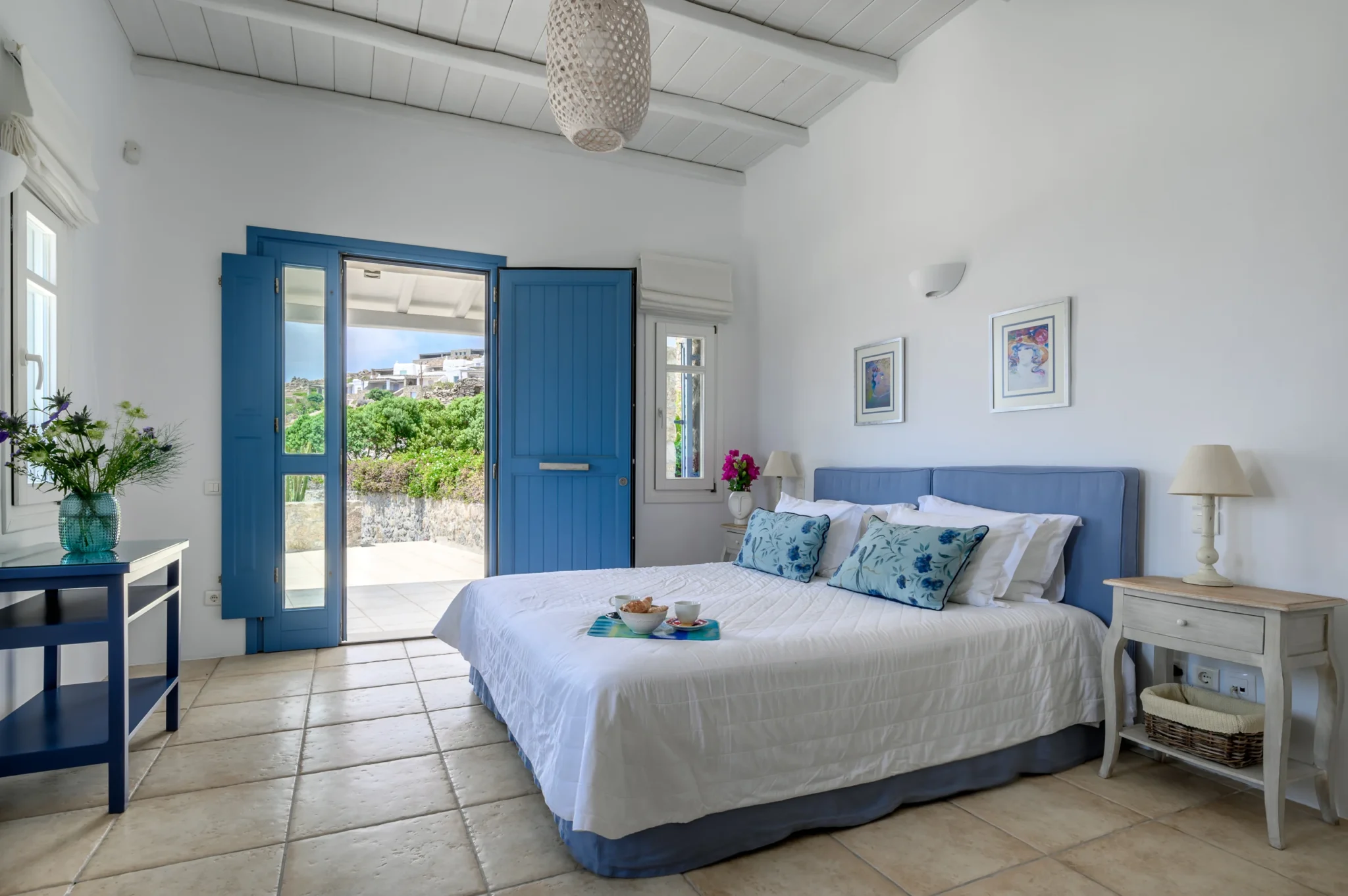 Bedroom with a blue headboard, floral cushions and breakfast tray, beside an open blue door to the terrace at Villa Mustique in Mykonos.